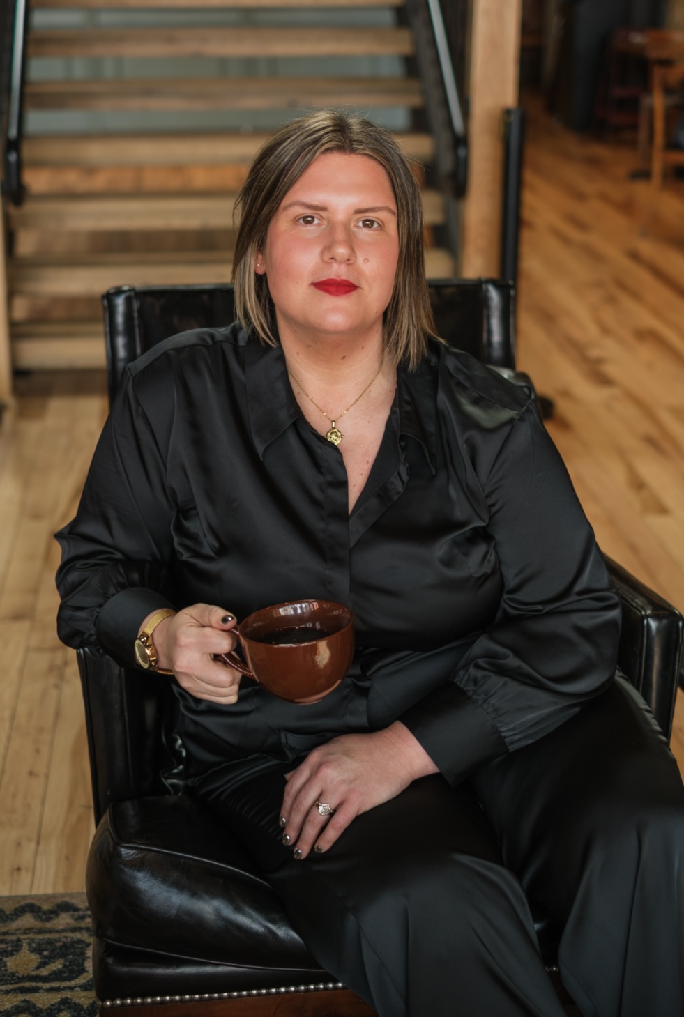Woman with shoulder-length hair and red lipstick sitting in a black chair, holding a brown mug, in a wooden interior.
