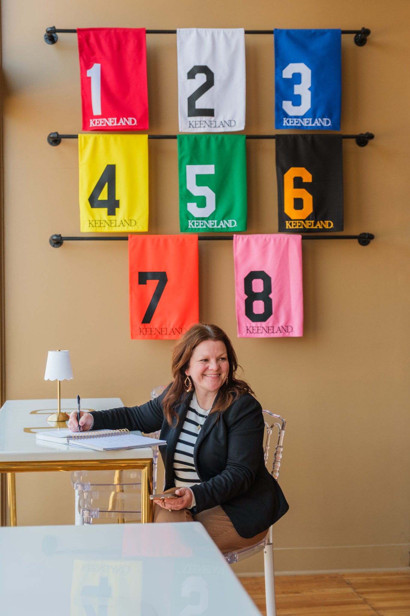 Woman sitting at a desk with colorful flags numbered 1 to 8 hanging on a wall behind her.