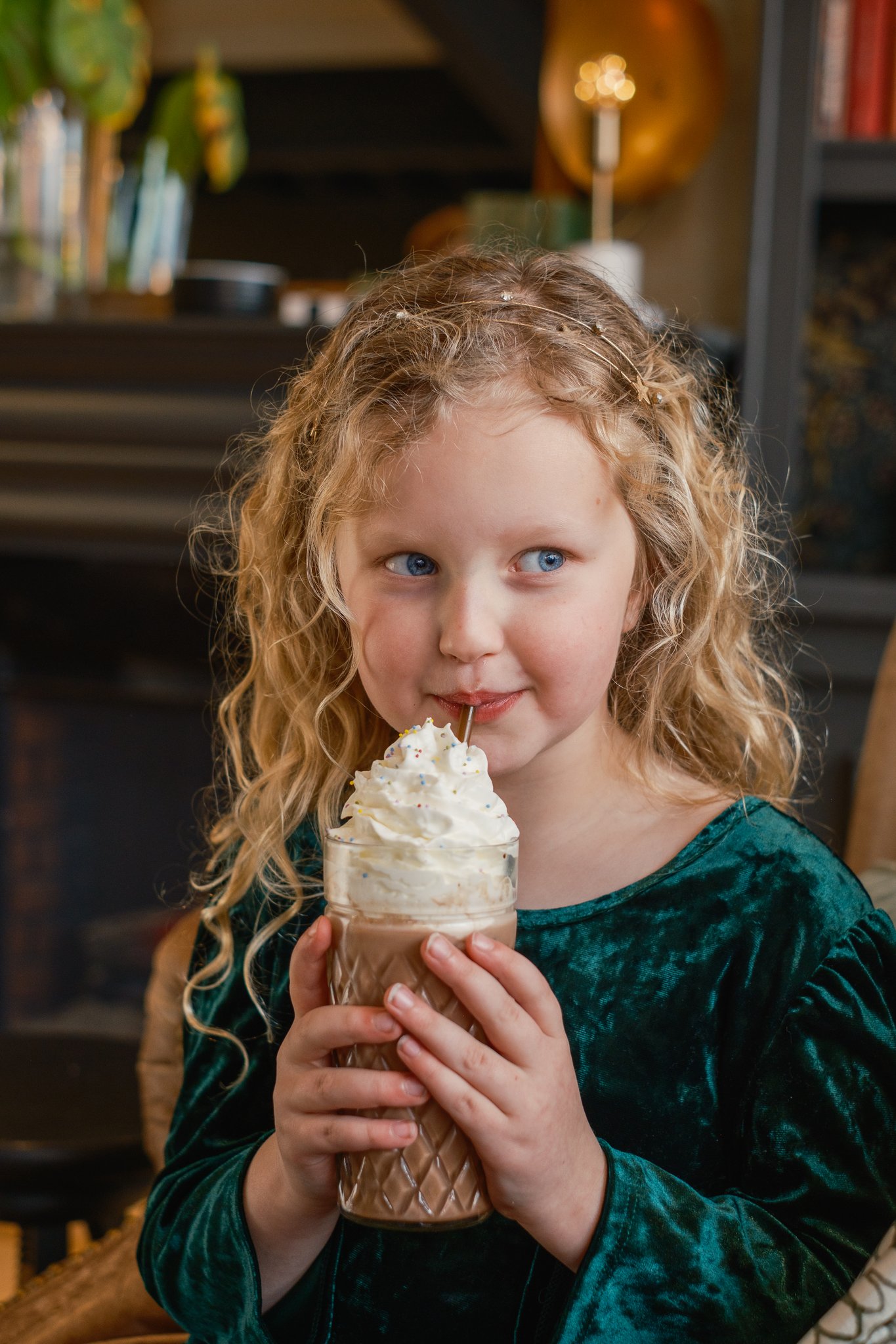 Young girl with curly blonde hair holding an ice cream cone, smiling, in a cozy indoor setting.