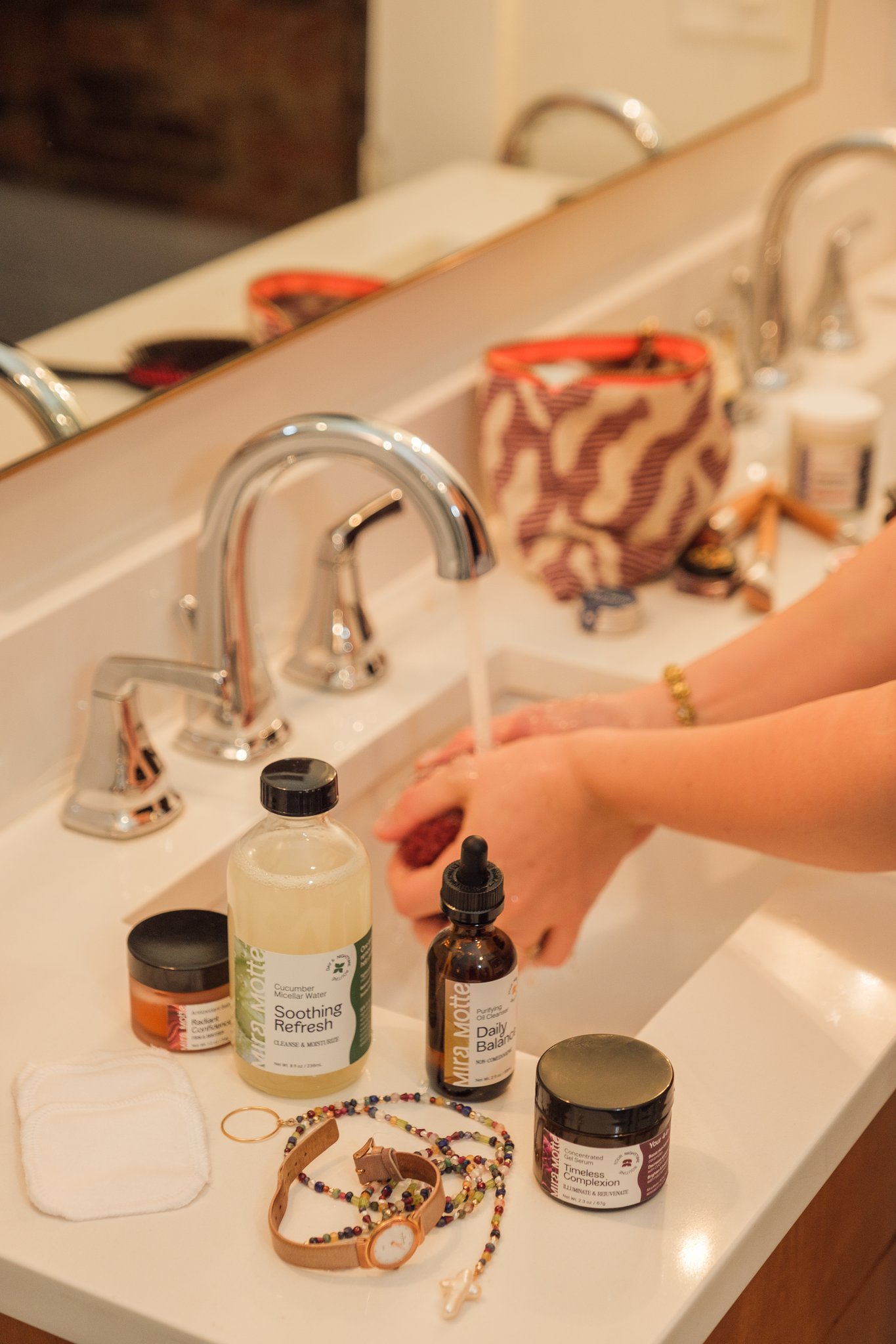 Person washing hands at bathroom sink with various skincare and jewelry items on the counter.
