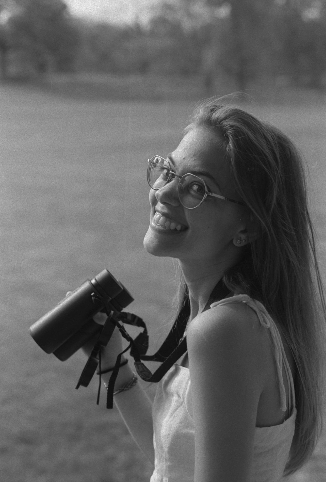 Smiling woman with glasses holding a camera outdoors, looking to the side, in a grassy area.