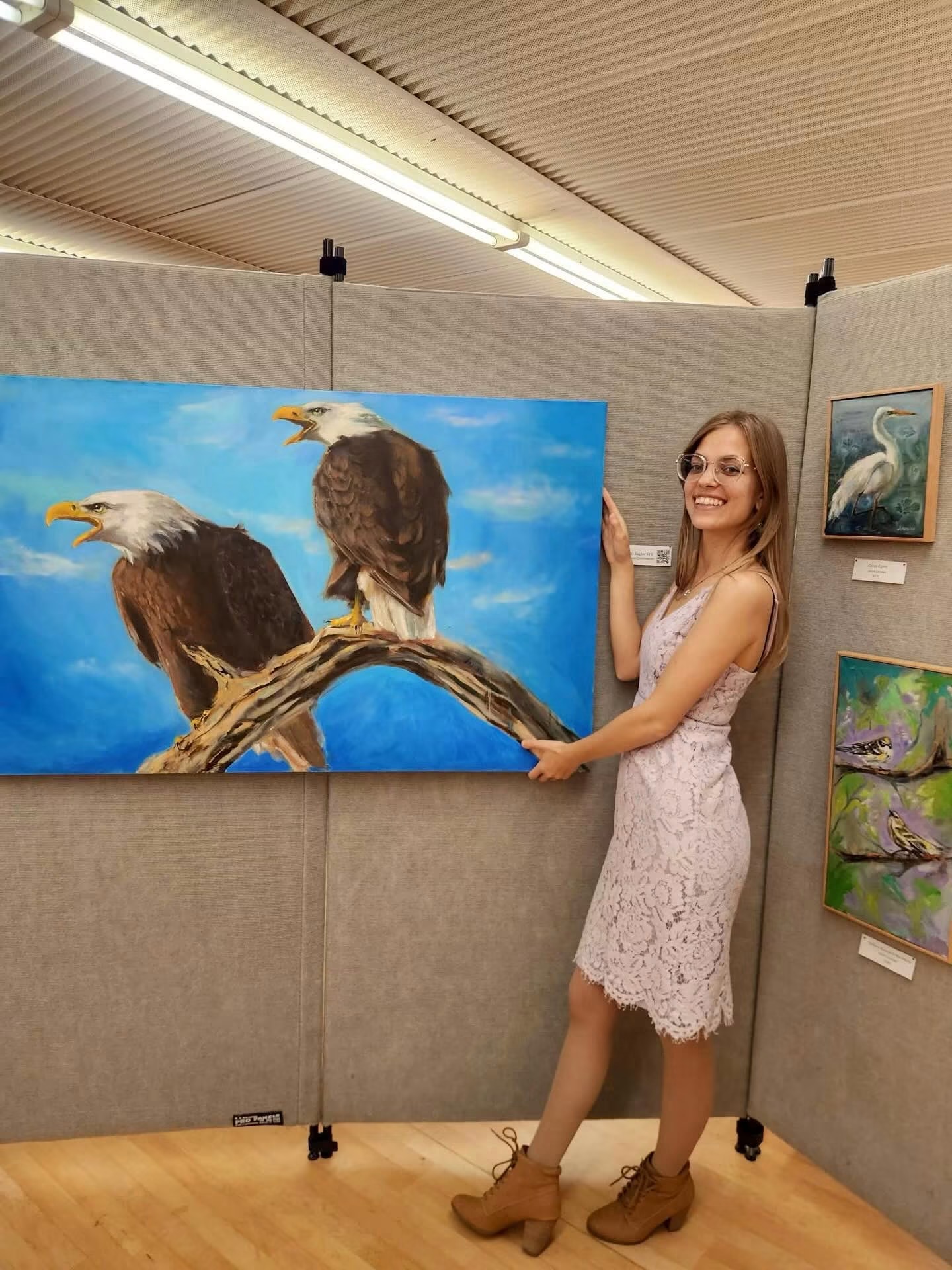 Young woman in a white lace dress and brown boots stands next to a painting of two eagles on a branch, smiling.