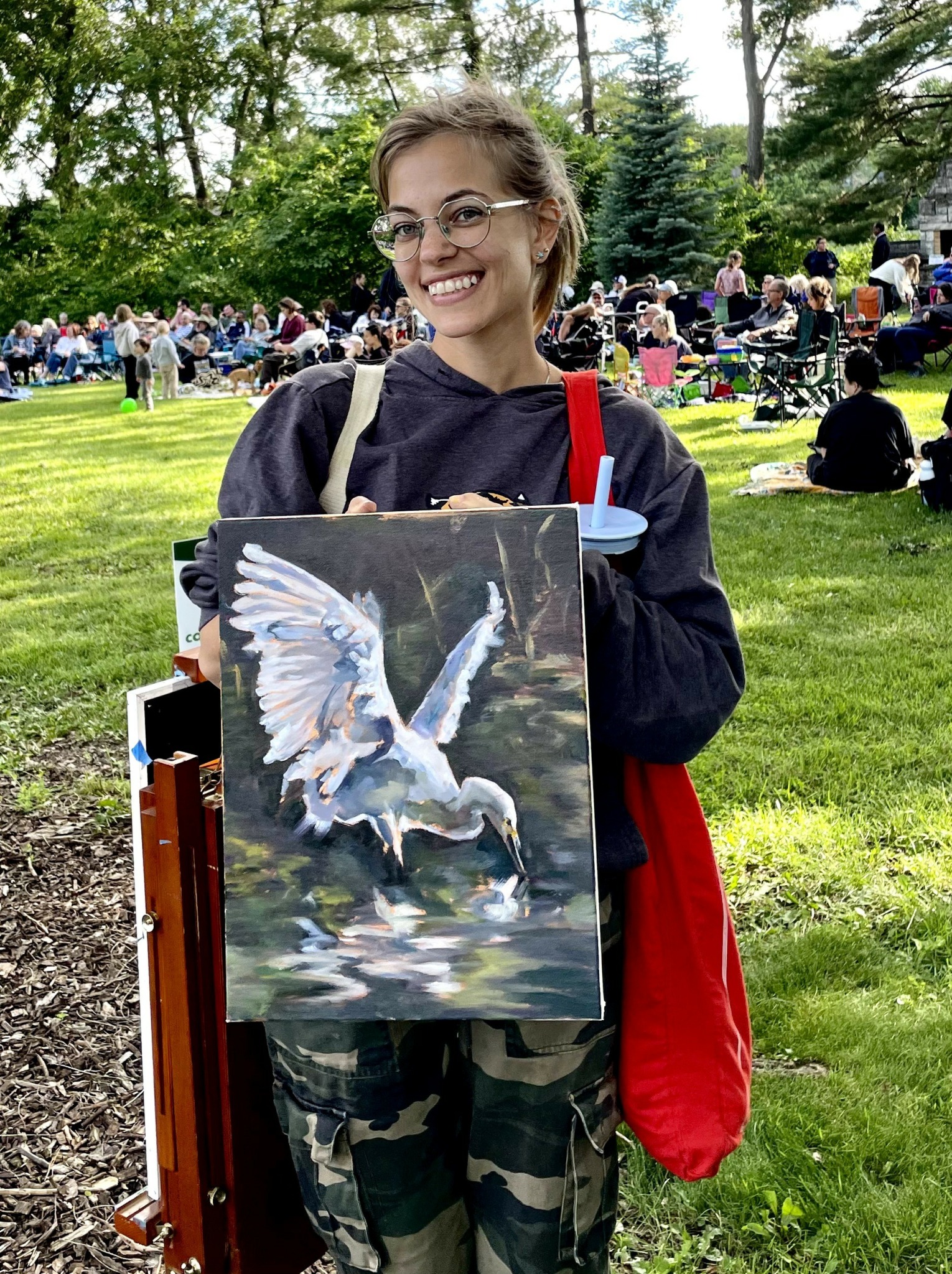 Young woman holding a painting of a white bird with wings spread, outdoors in a park with people and trees.