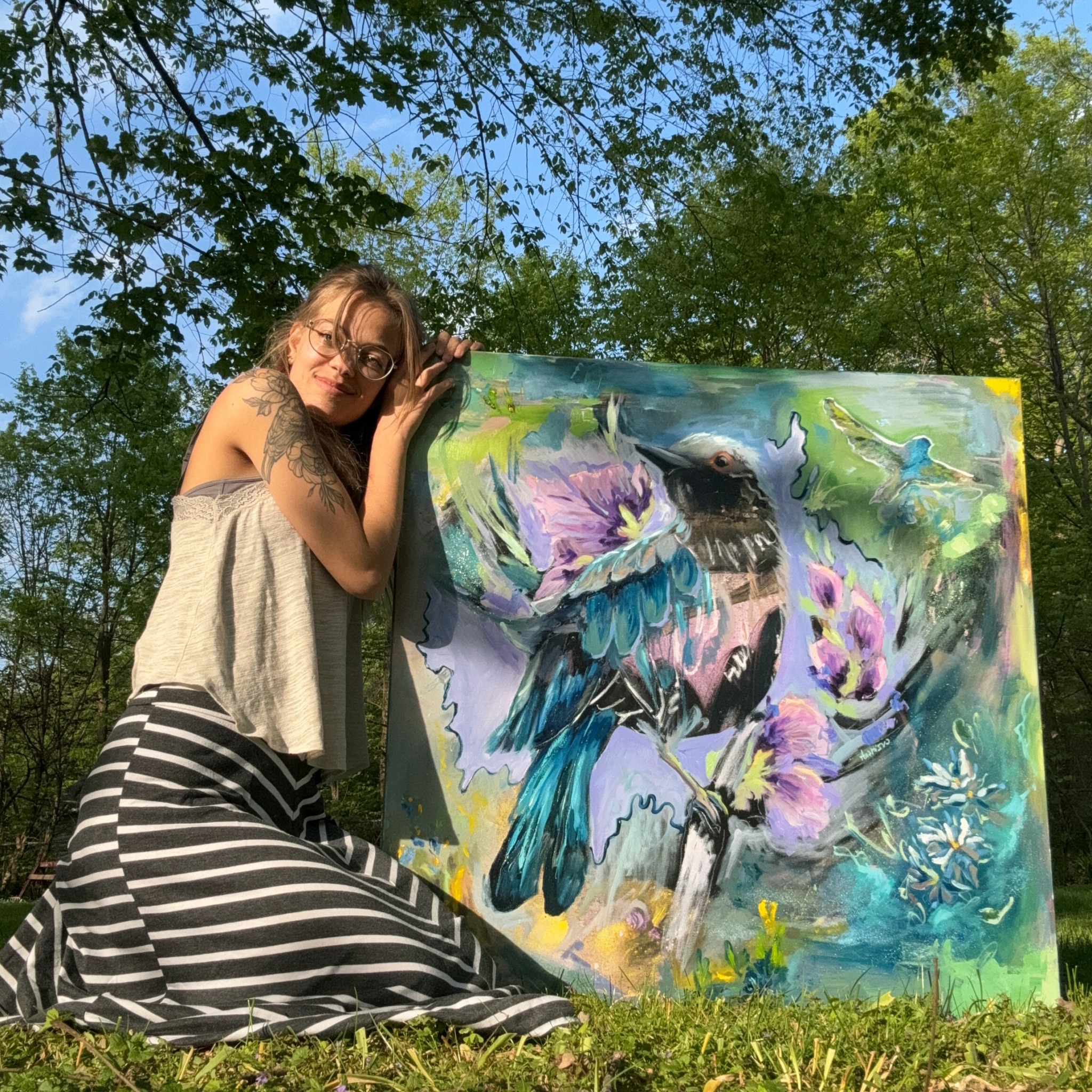 Woman with glasses and long hair leaning on colorful bird painting outdoors, surrounded by trees and blue sky.