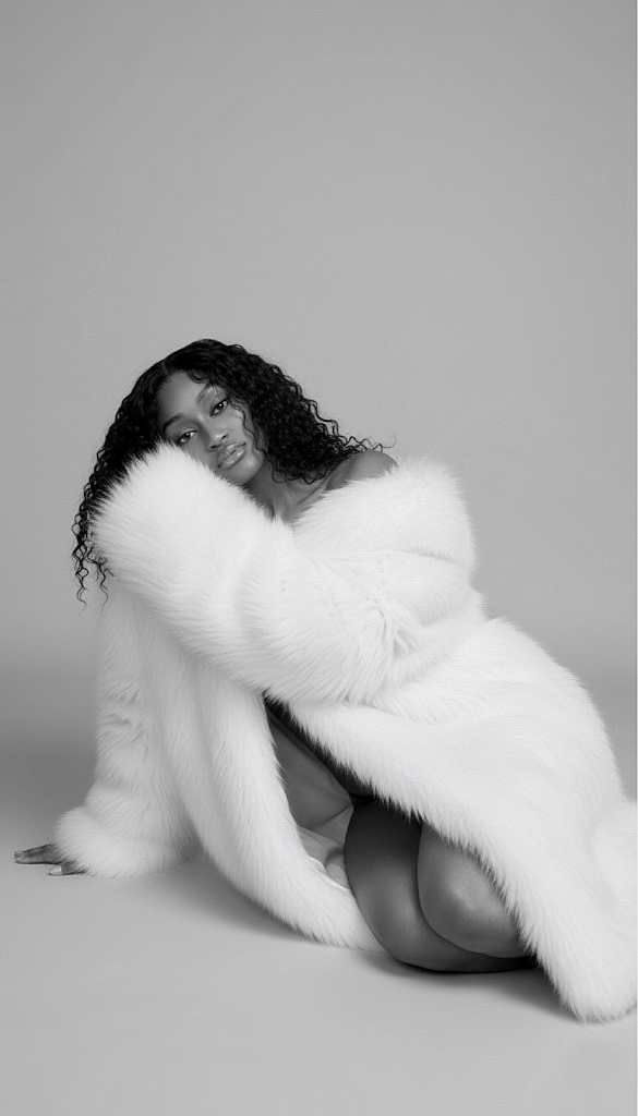Woman with curly hair wearing a white fur coat, sitting on the floor against a plain background.