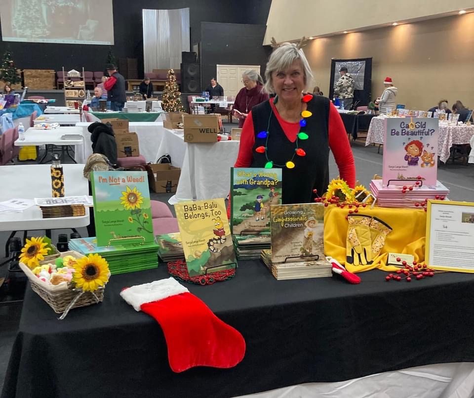 Woman standing behind a table with books, sunflowers, a Christmas stocking, and holiday decorations at an indoor event.