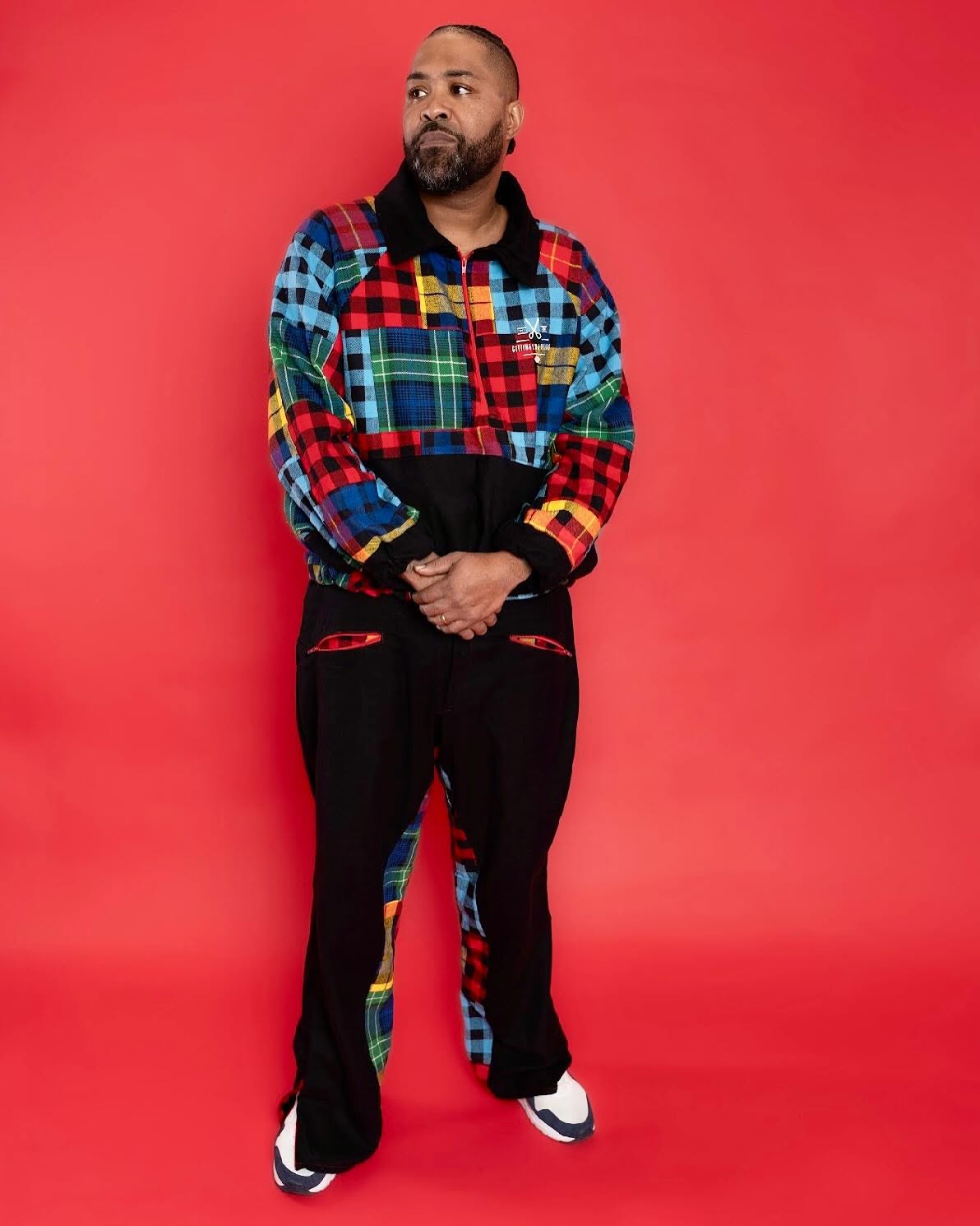 Man with a beard in colorful patchwork jacket and black pants stands against red background.