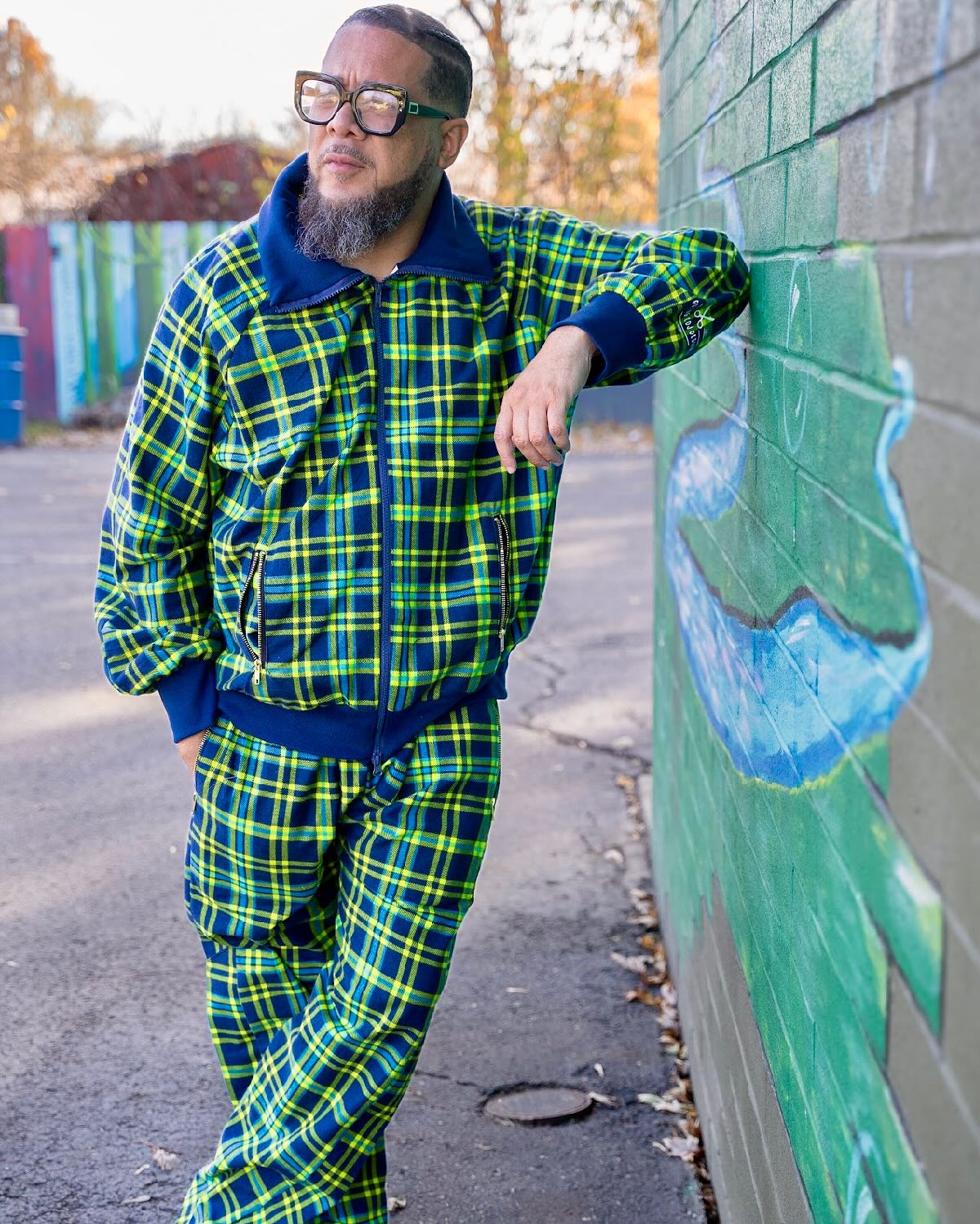 Man with glasses and beard leaning against a wall outdoors, wearing a green and blue plaid outfit, with trees and a colorful fence in background.