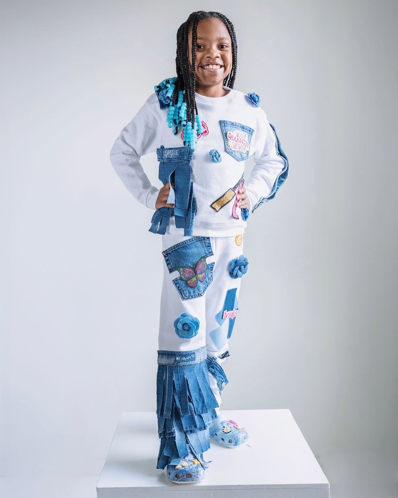 Child with braided hair wearing a white outfit decorated with blue patches, fringe, and accessories, standing on a white platform.