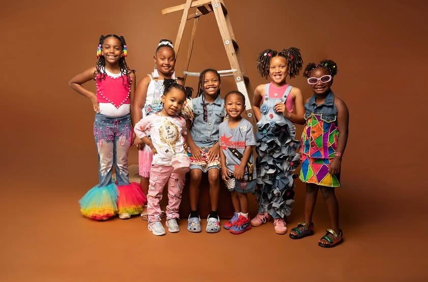 Group of eight children standing in front of a brown background, some wearing colorful outfits and accessories, smiling.