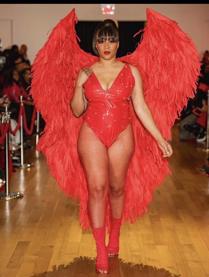 Model walking runway in red outfit with large feathered wings, fishnet stockings, and high heels, audience watching in background.