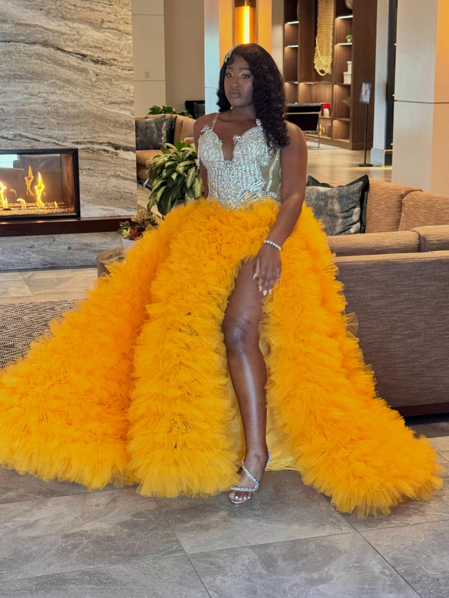 Woman in a yellow feathered gown with a high slit, standing indoors near a fireplace and sofa.