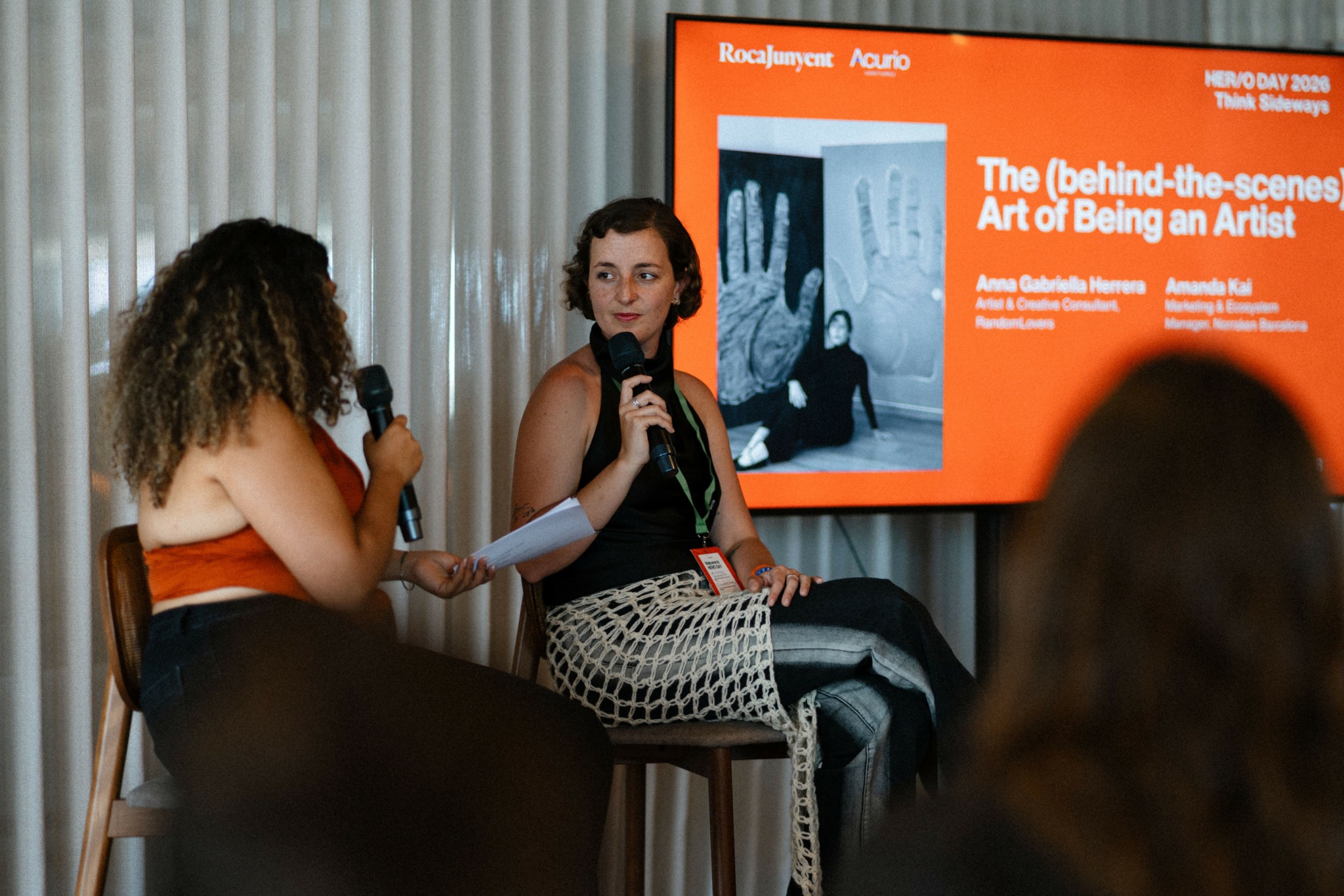 Two women sit on chairs, one speaking into a microphone, with a presentation slide behind them. Audience members are visible.