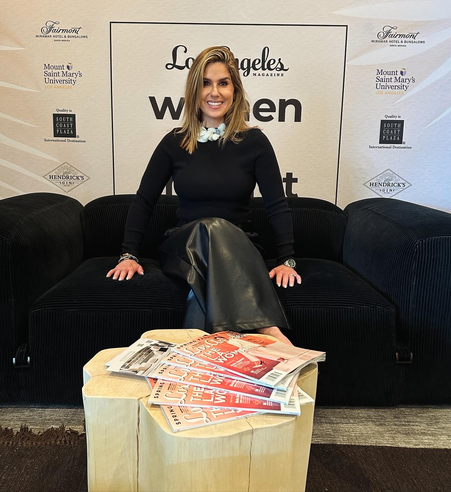 Woman sitting on a black sofa with a backdrop behind her, a table with newspapers and magazines in front, smiling.
