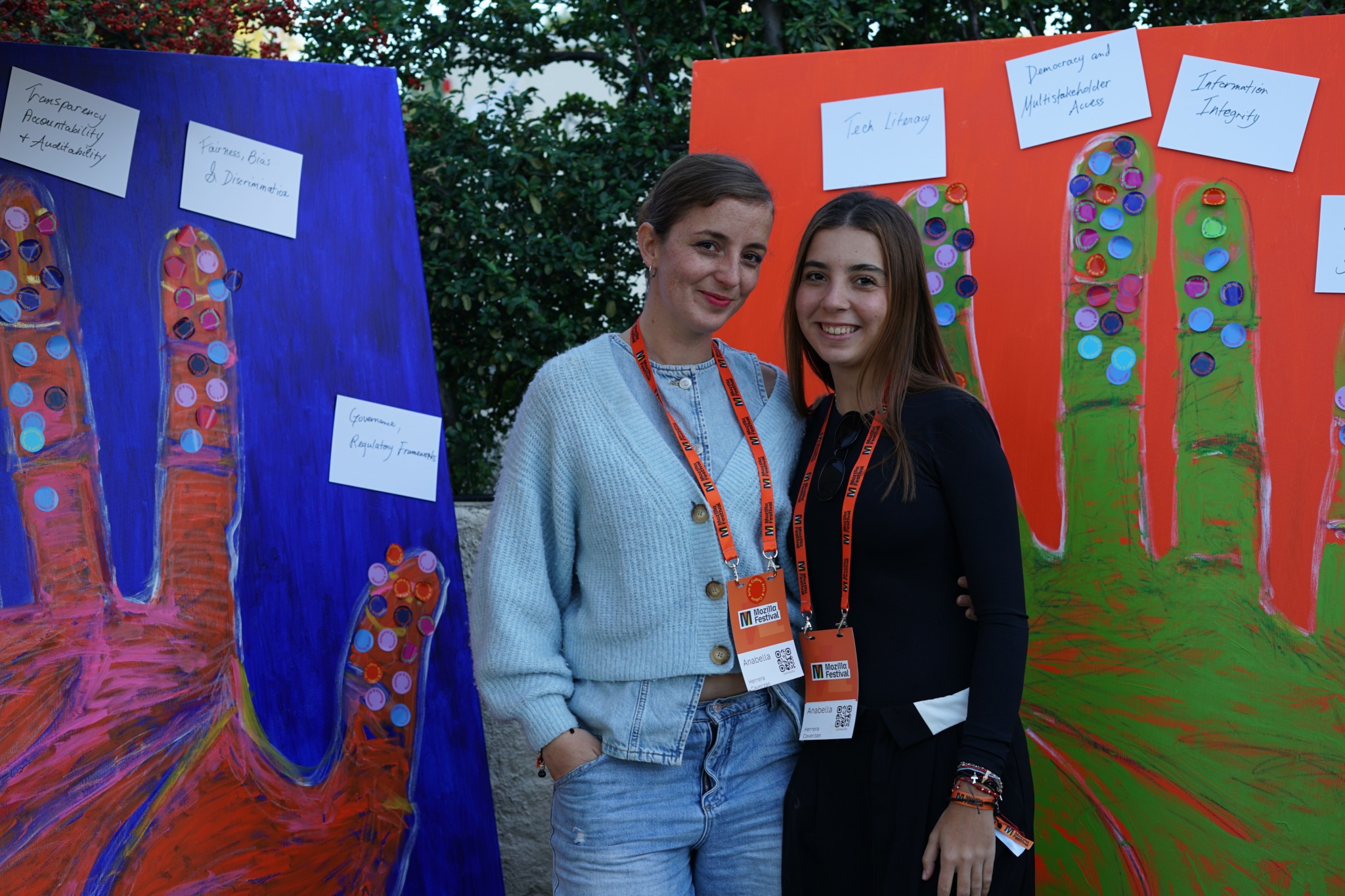 Two women standing outdoors in front of colorful display boards with trees and notes, smiling at the camera.