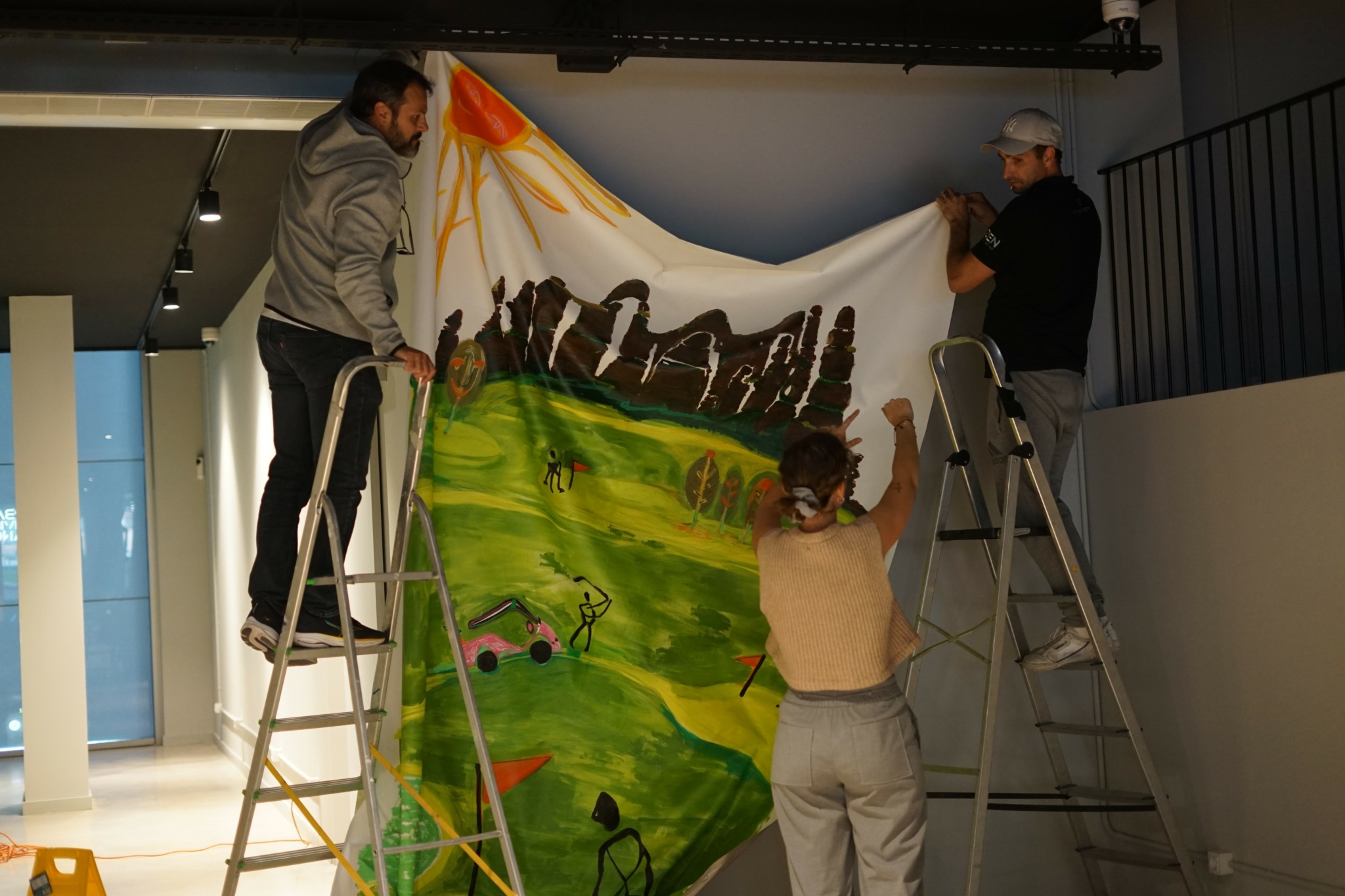 Three people hang a large colorful fabric mural depicting mountains, green fields, and a bright sky indoors.
