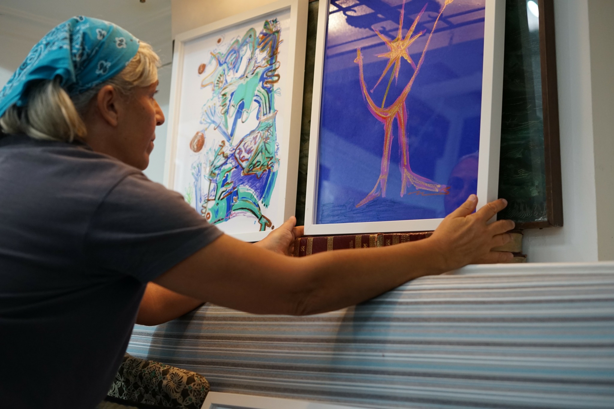 Person with a blue headscarf adjusting a colorful abstract artwork on a window ledge.