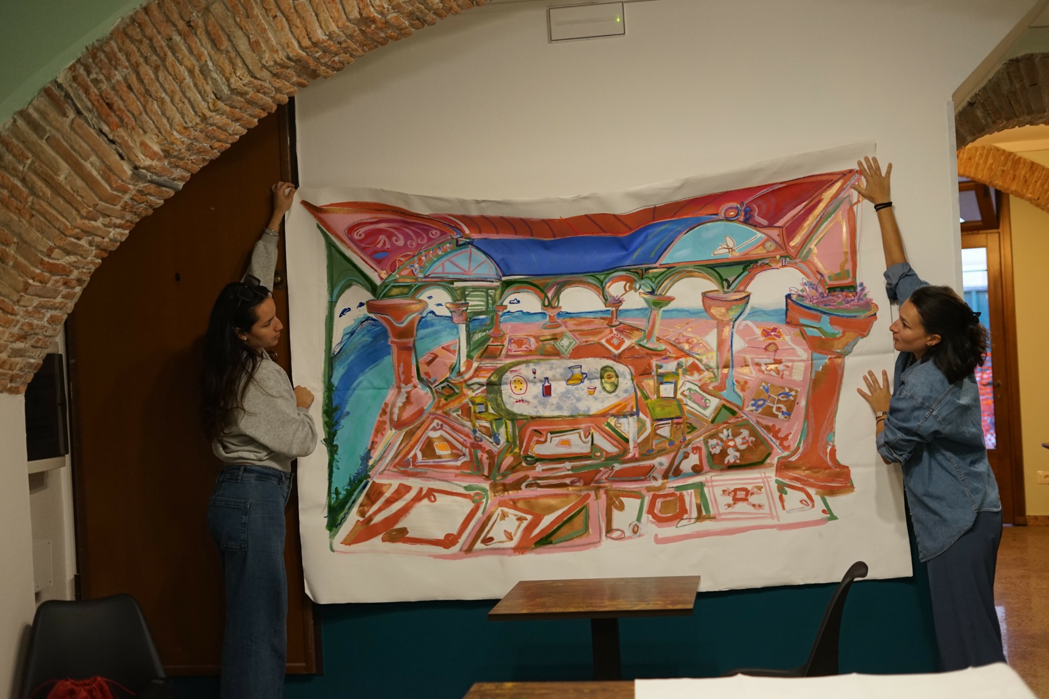 Two women stand on either side of a colorful, abstract painting hung on a white wall, with brick arches overhead.