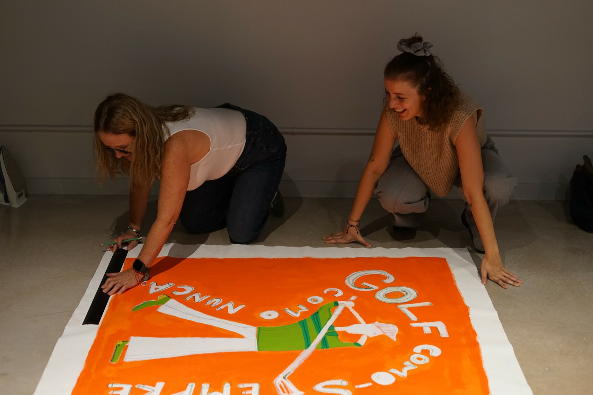 Two women are kneeling on the floor, working on a large orange and white poster with a golf-themed design.