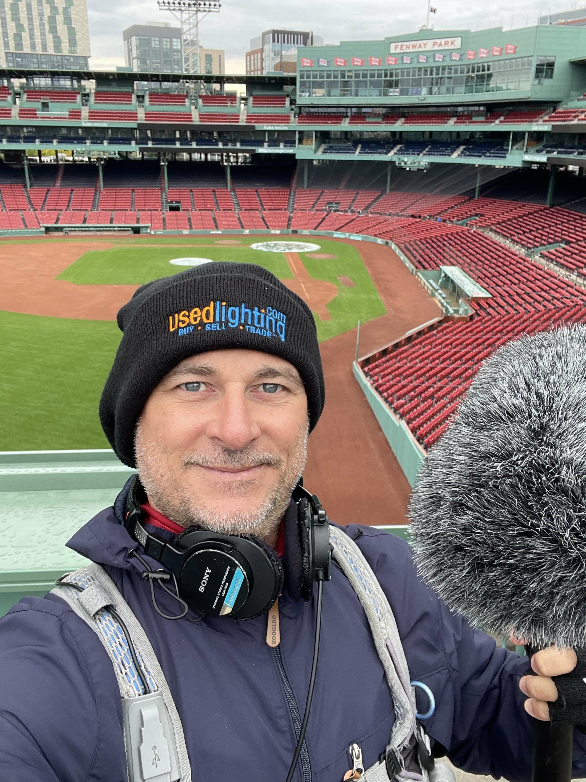 Man wearing a black beanie and headphones at a baseball stadium, with empty red seats and a green field in the background.