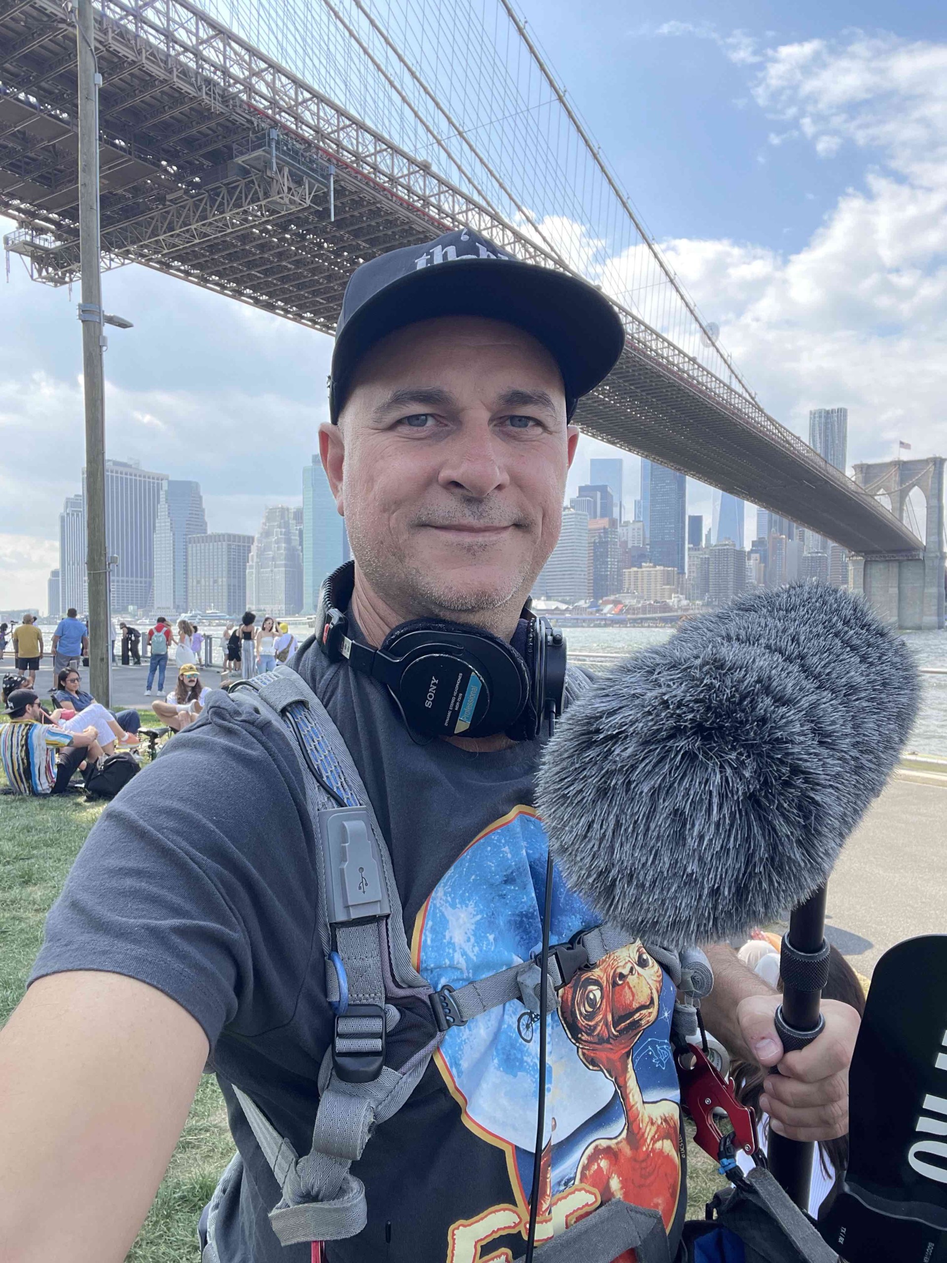 Man taking selfie outdoors with city skyline and bridge in background, holding a microphone with a furry windscreen, wearing a cap and headphones.