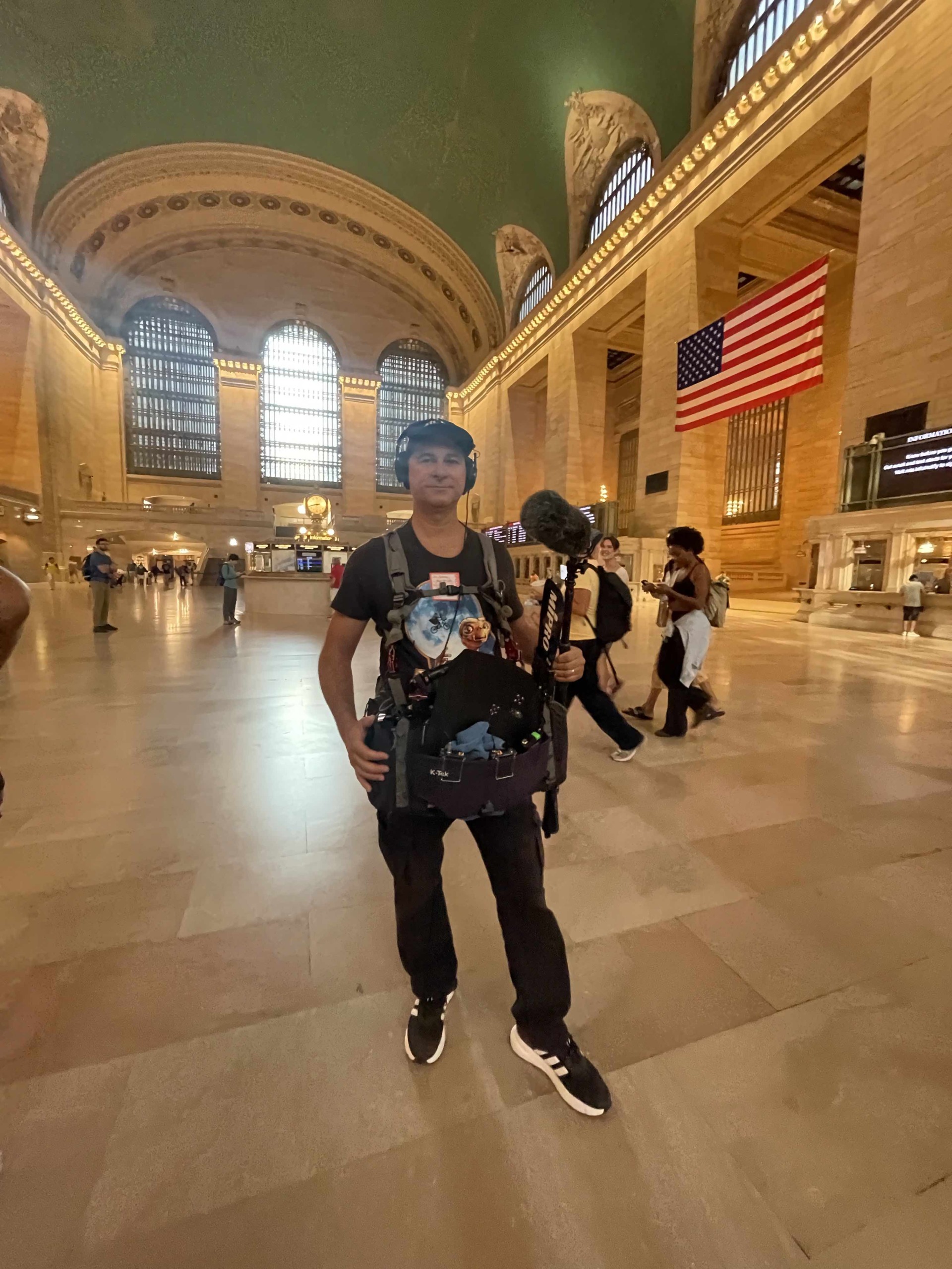 Person standing inside a grand train station with high arched ceiling and American flag, carrying a backpack.
