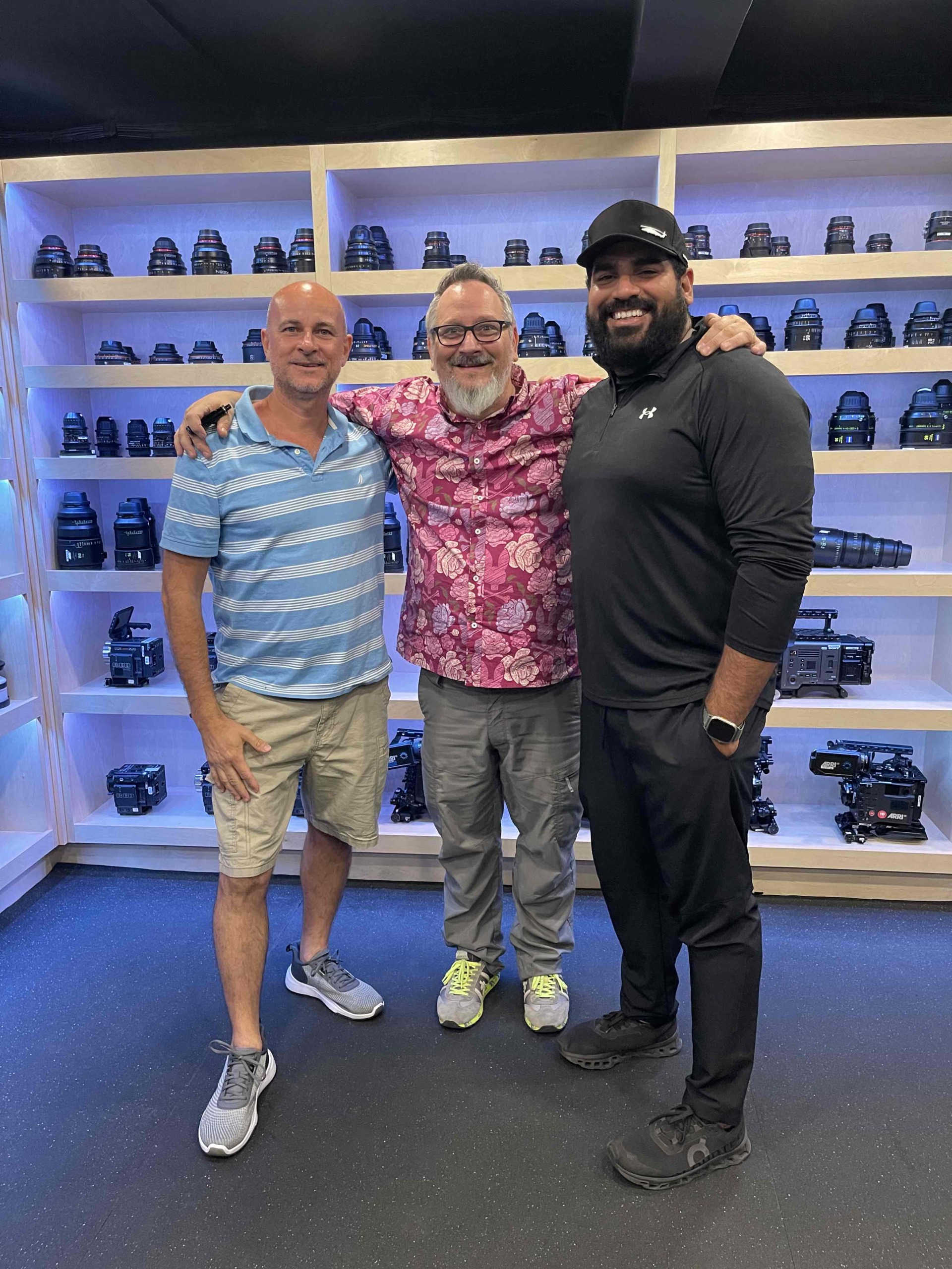 Three men standing together in front of shelves filled with cameras, smiling and posing for the photo.