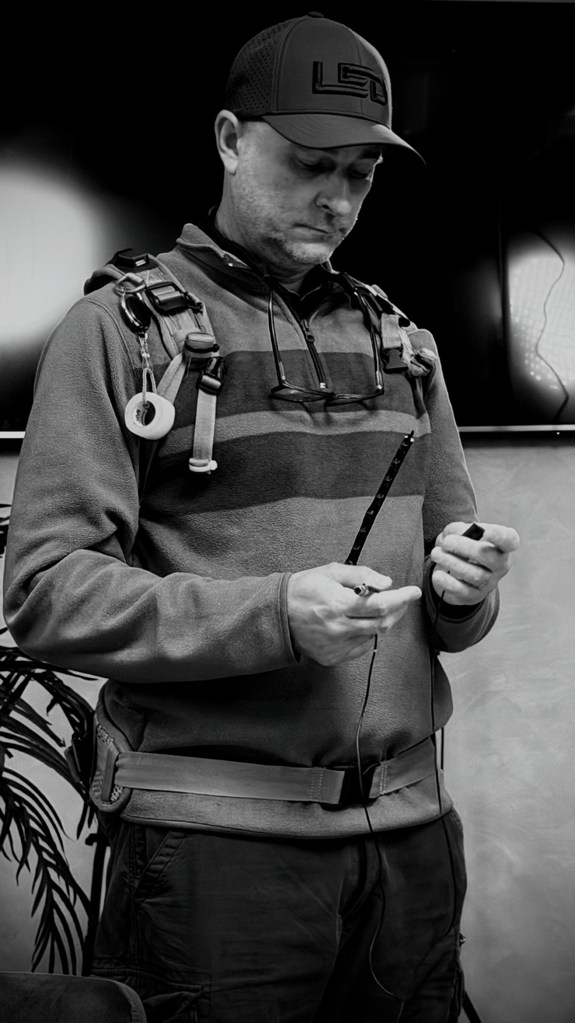 Young man in cap and jacket looking at a device, standing indoors with a plant nearby.