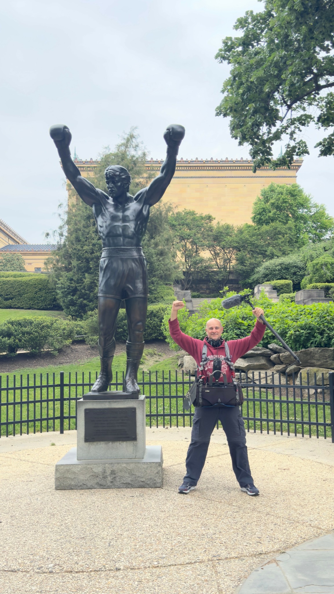 Person standing with arms raised in front of a large statue of a muscular man with arms raised, outdoors with trees and a building.