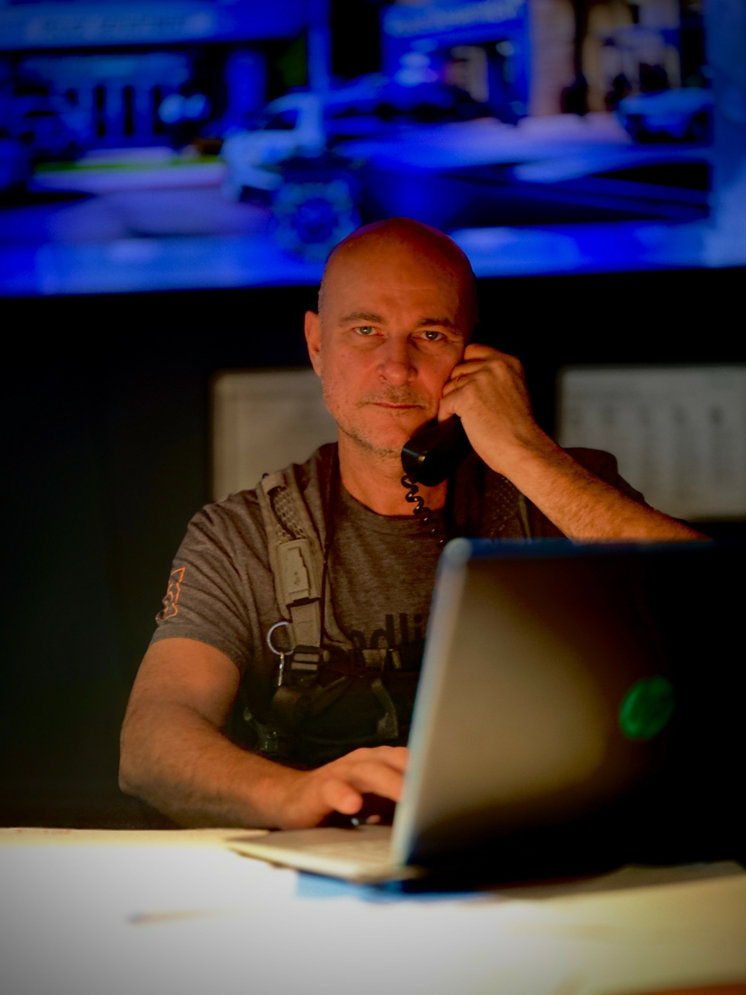 Man using a laptop and talking on a phone in a dimly lit room with a screen displaying a cityscape in the background.