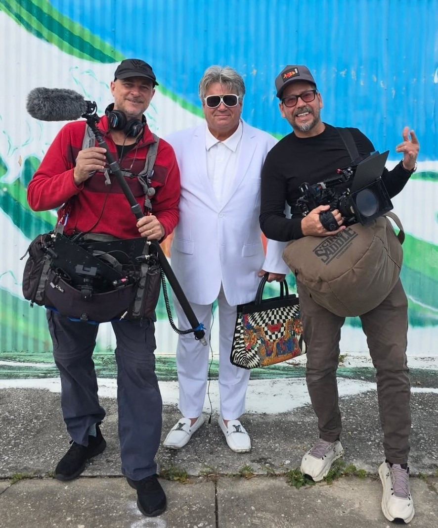 Three men standing outdoors in front of a colorful background, two holding camera equipment, one in white suit, all smiling.
