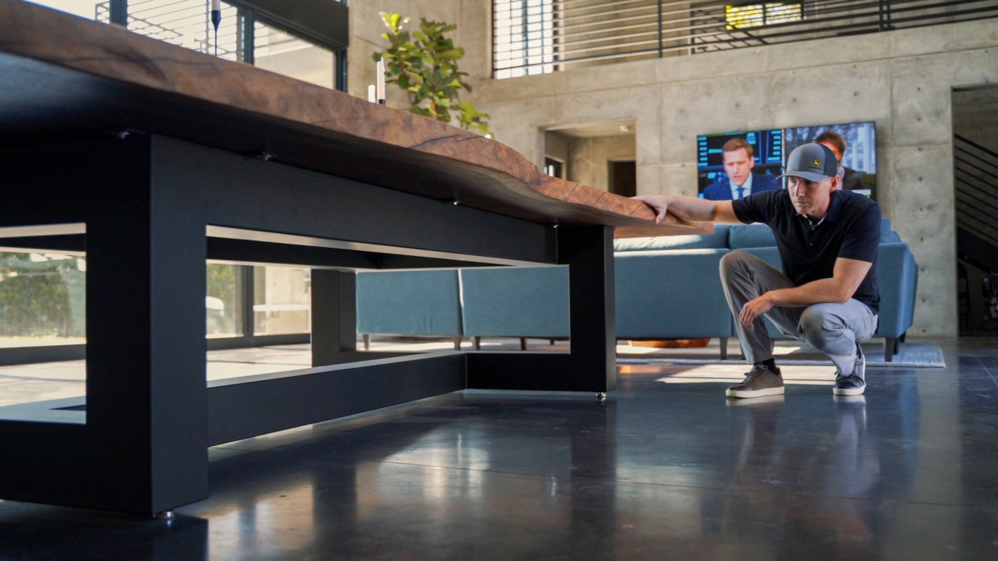 Man crouching near a long table in a modern indoor space with large windows and a television screen in the background.