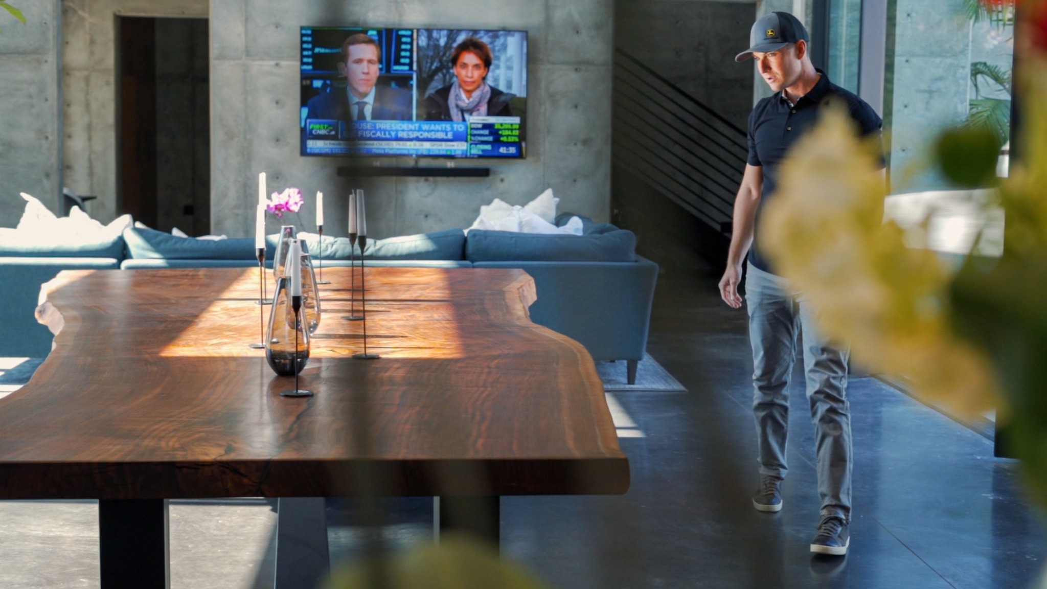 Modern living room with a wooden table, TV on the wall, and a person walking nearby, blurred flowers in foreground.