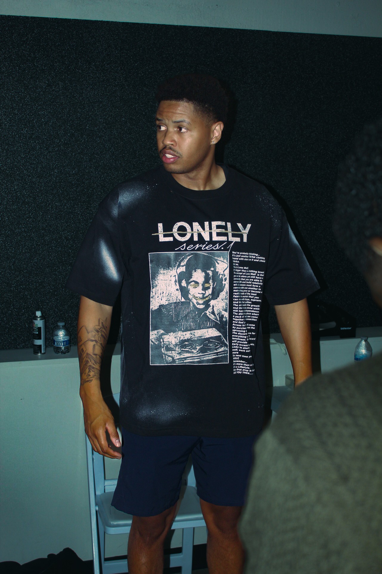 Young man with short hair wearing a black graphic T-shirt and dark shorts, standing indoors against a dark background.
