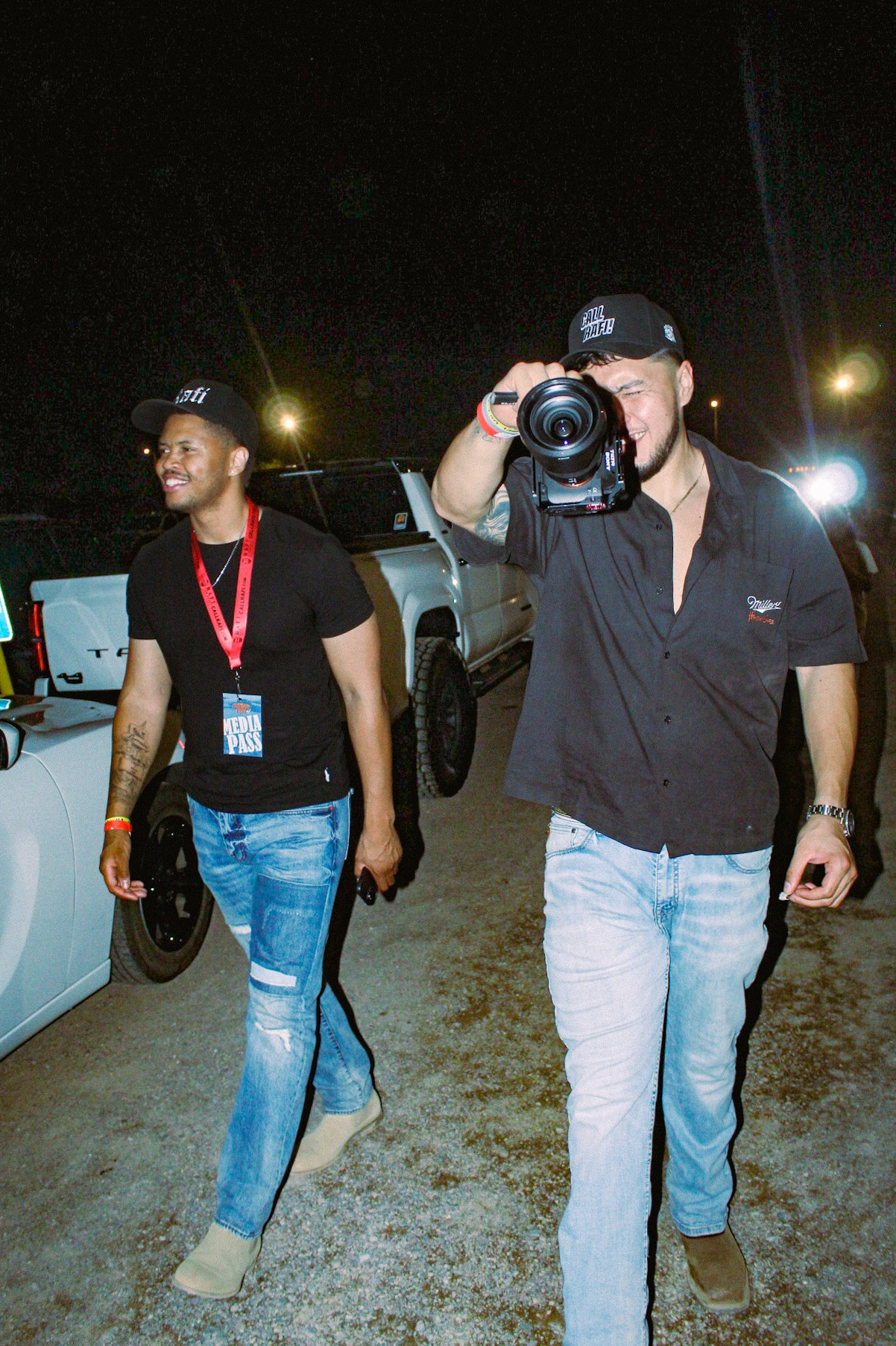 Two men walking outdoors at night, one taking a photo with a camera, both wearing black caps and casual clothing.