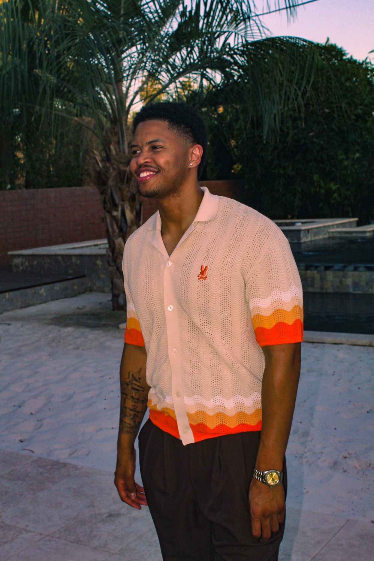 Young man smiling outdoors with palm trees and a fence in the background, wearing a light-colored shirt with orange accents.