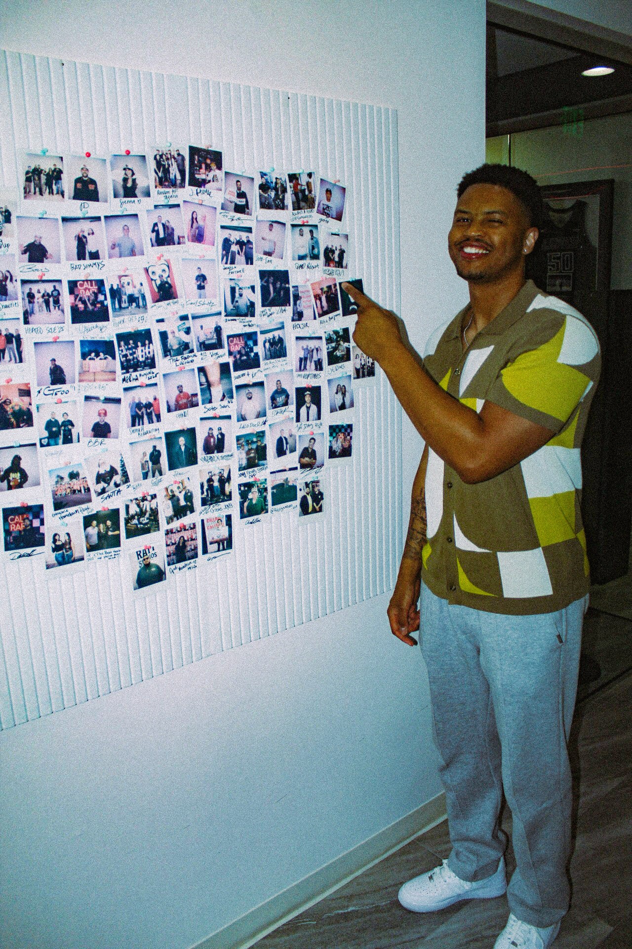 Person smiling and pointing at a wall display of numerous photographs, standing indoors with a dark ceiling and white wall.