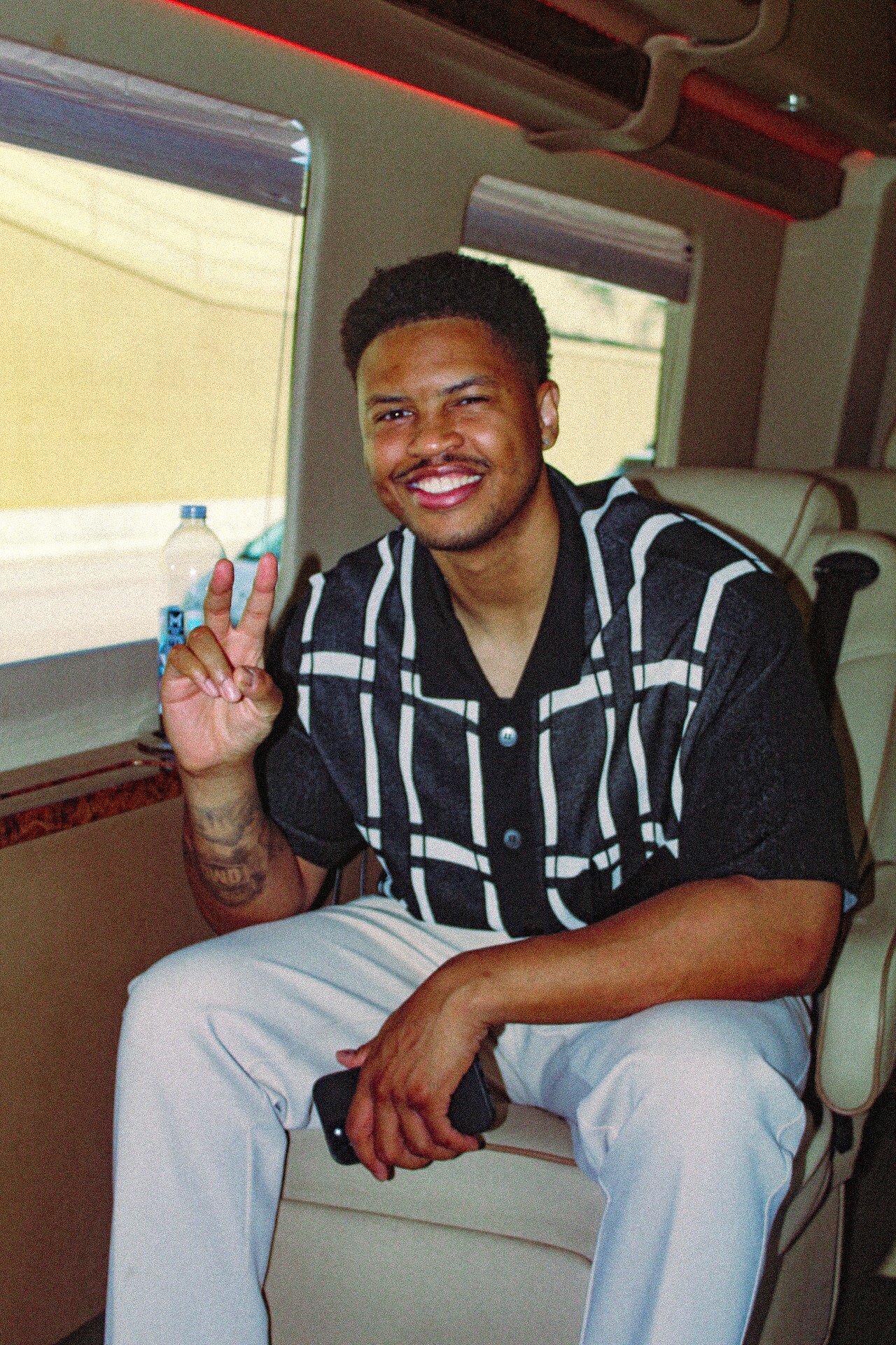 Young man with short curly hair and a beard, smiling, sitting on a bus seat, wearing a black and white striped shirt, holding up a peace sign with his right hand.