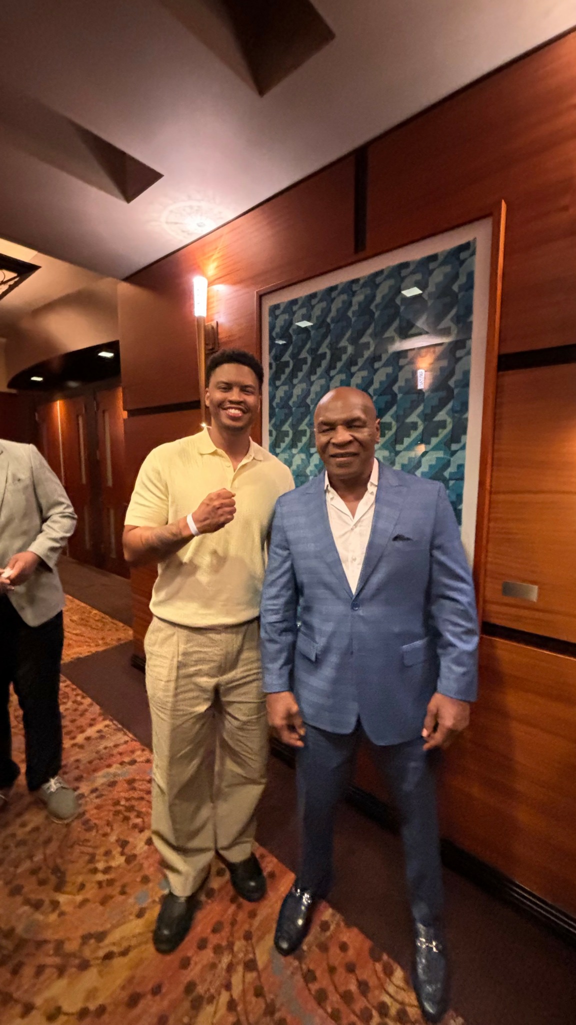Two men standing indoors, one in a light yellow shirt and beige pants, the other in a blue suit, smiling.