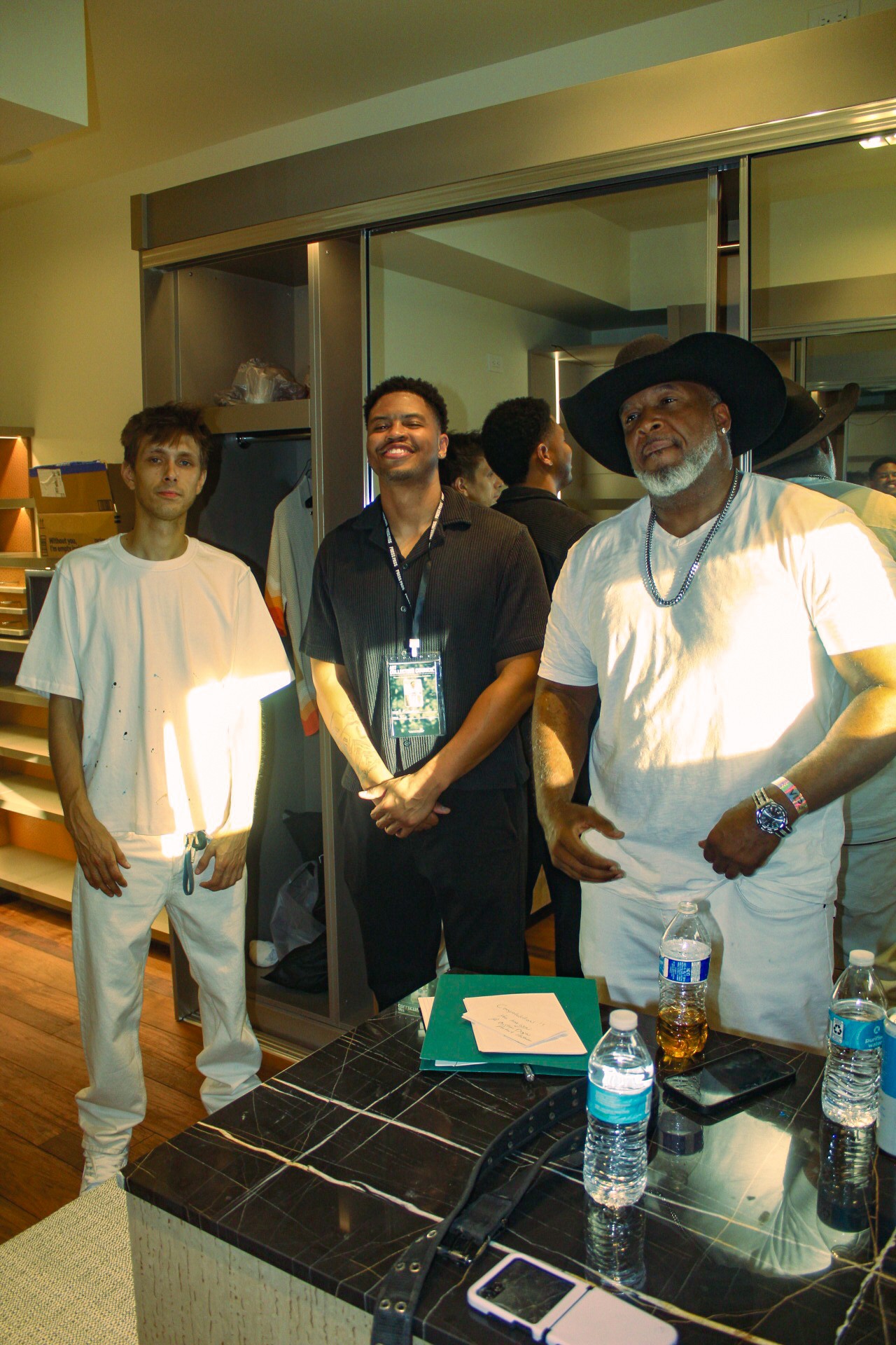 Three people standing indoors near a table with water bottles and papers, smiling at the camera.