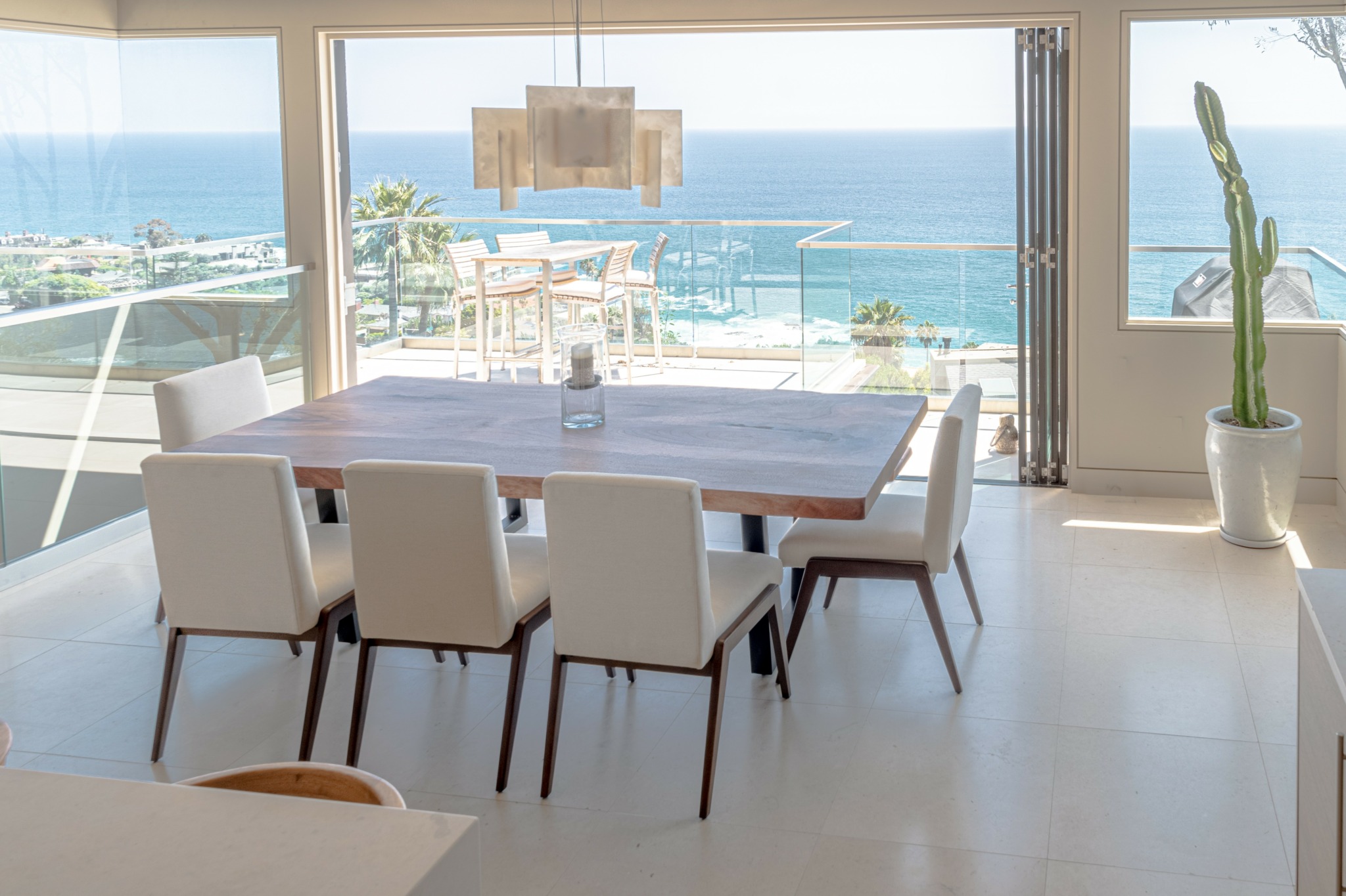 Dining room with a large table and six chairs, ocean view through large windows, potted cactus, and a hanging light fixture.