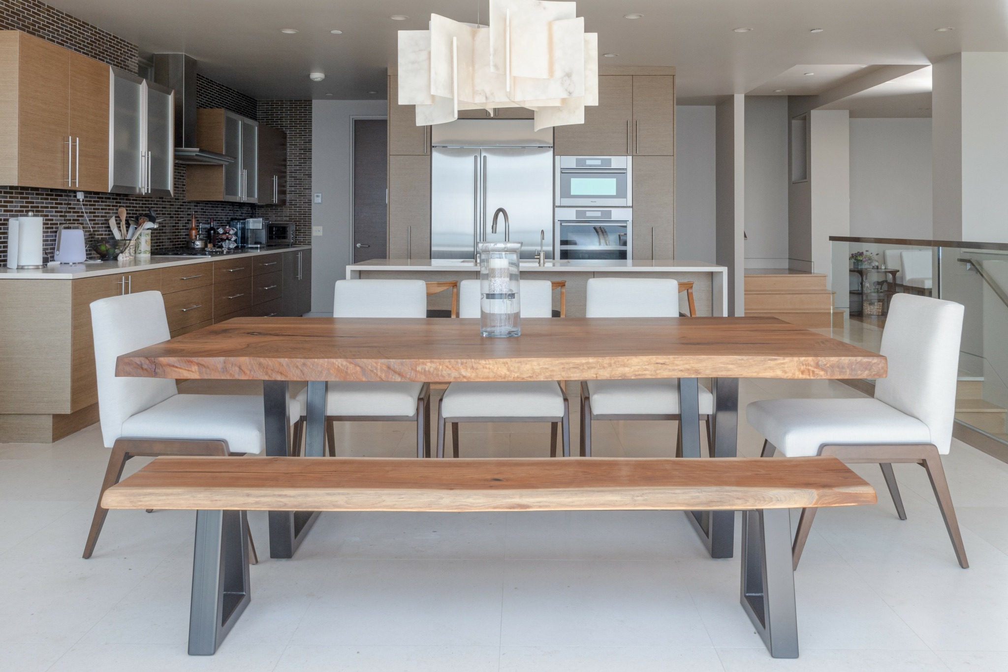 Modern kitchen and dining area with a wooden table, white chairs, and a bench, illuminated by a large ceiling light.
