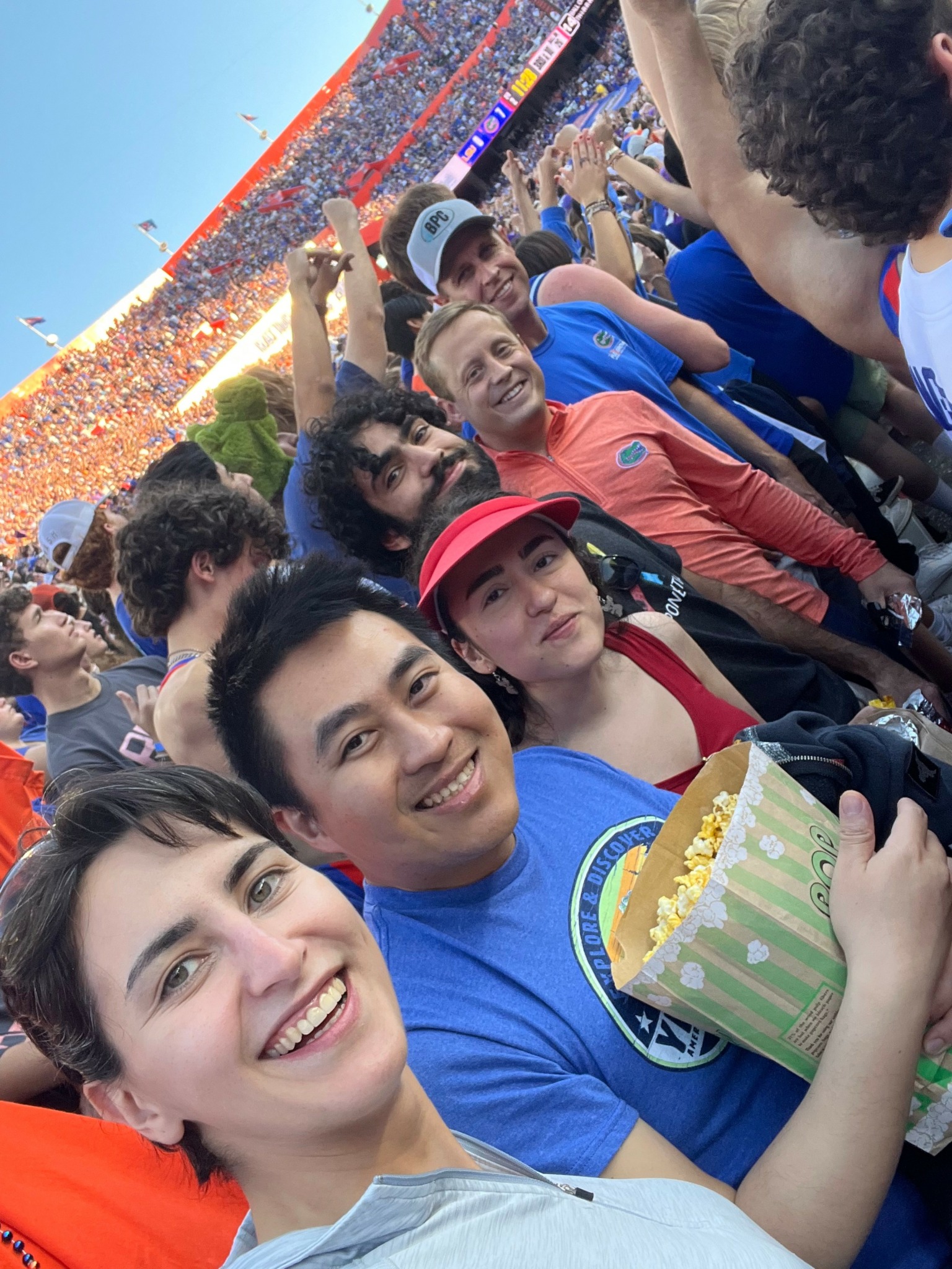 Group of people smiling at a stadium event, some holding popcorn, with a large crowd and stadium in background.
