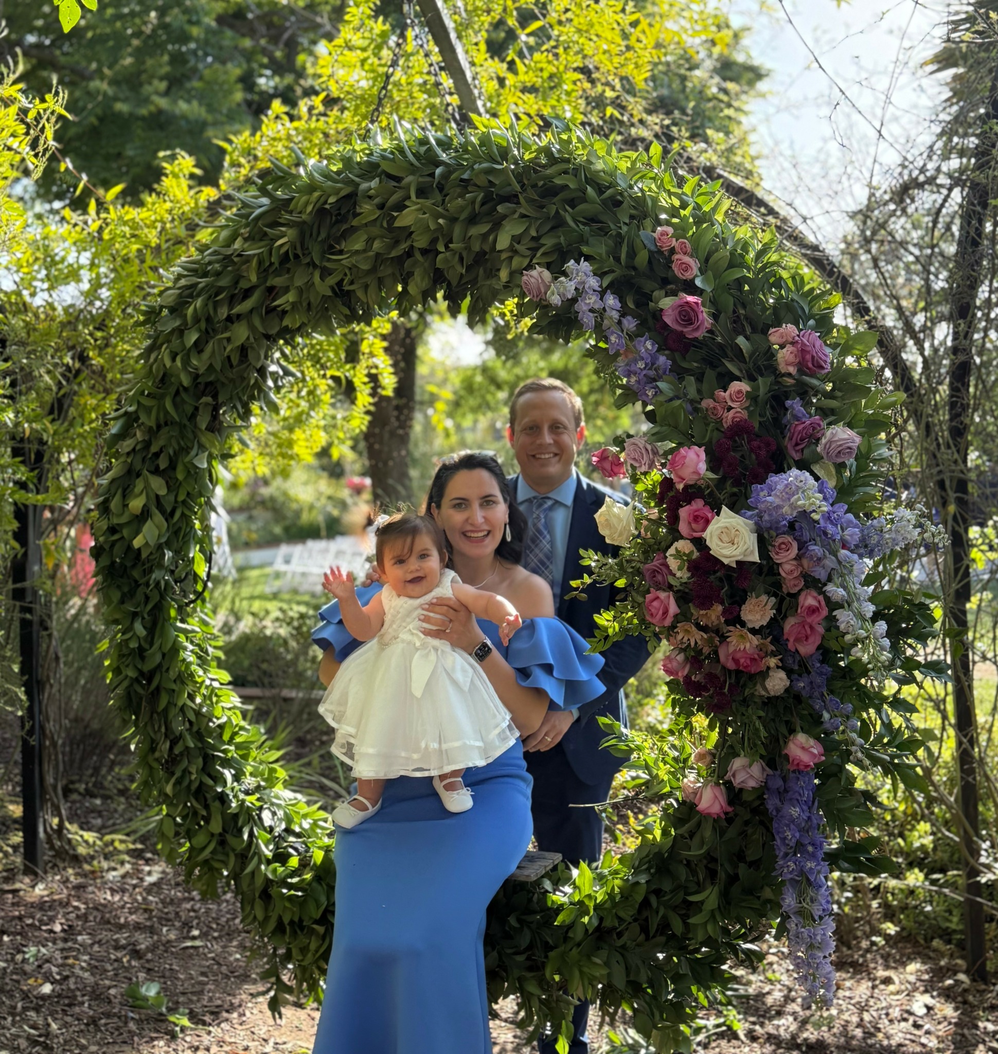 A family of three stands inside a floral heart-shaped arch outdoors, smiling and posing for a photo.