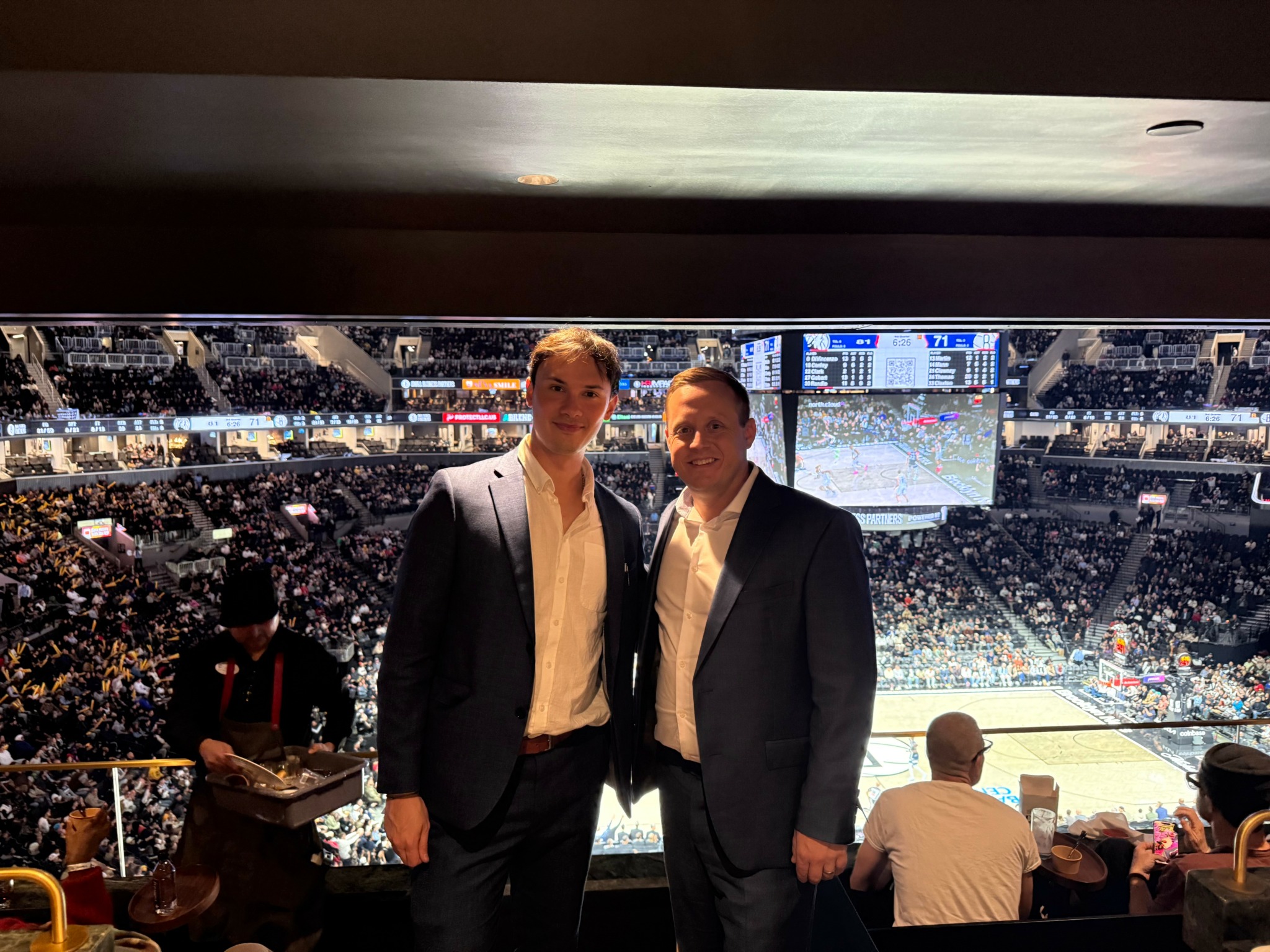 Two men in suits standing in a sports arena with a large crowd and scoreboard in the background.