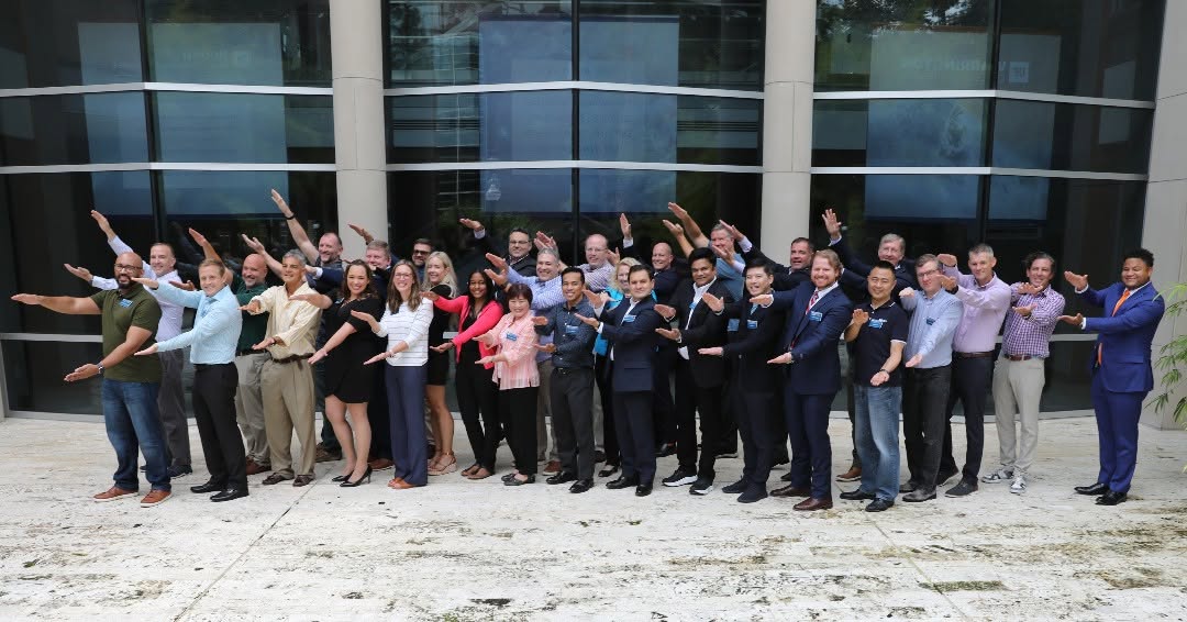Group of people standing outdoors in front of a building, posing with arms extended outward, smiling.