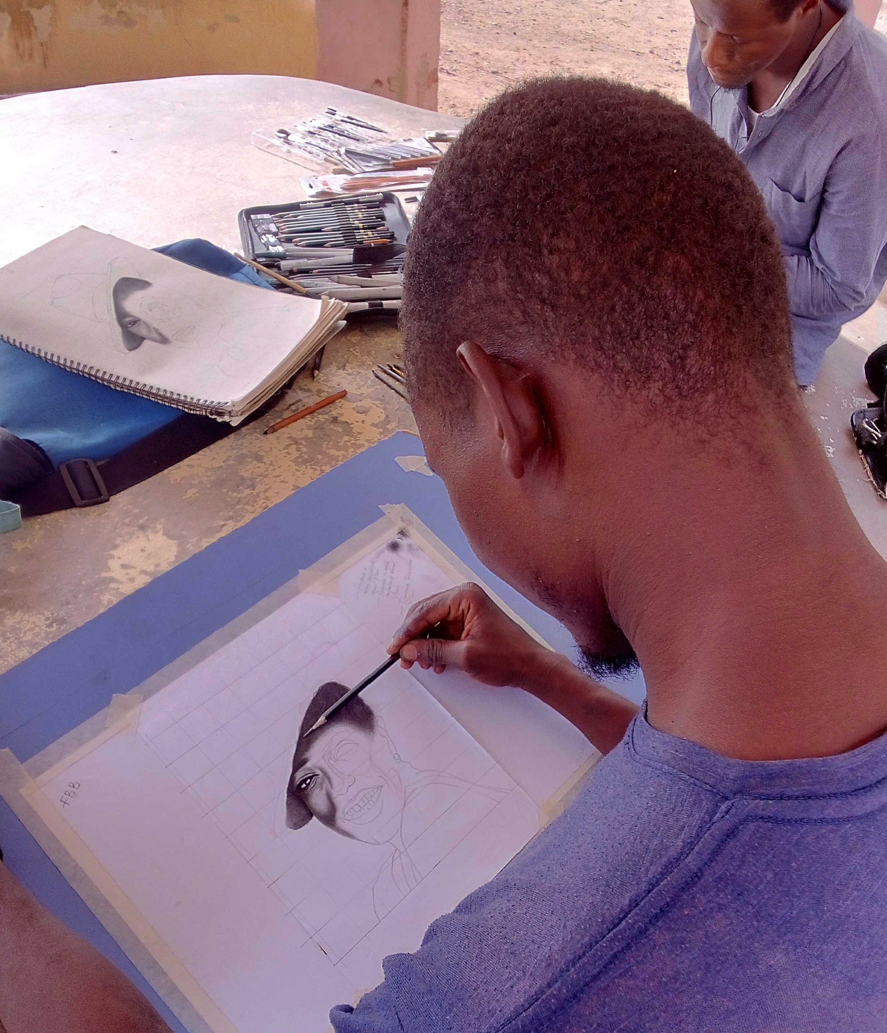 Young man drawing in a sketchbook at a table outdoors, with notebooks and supplies nearby.