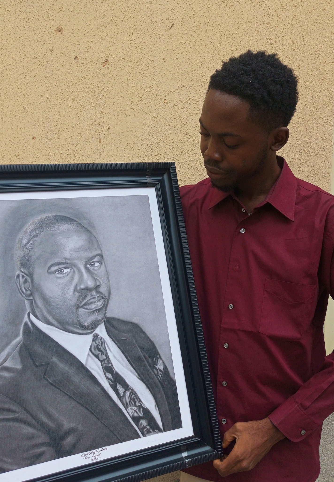 Young man with short curly hair in a maroon shirt holding a framed black-and-white portrait of a man in a suit.
