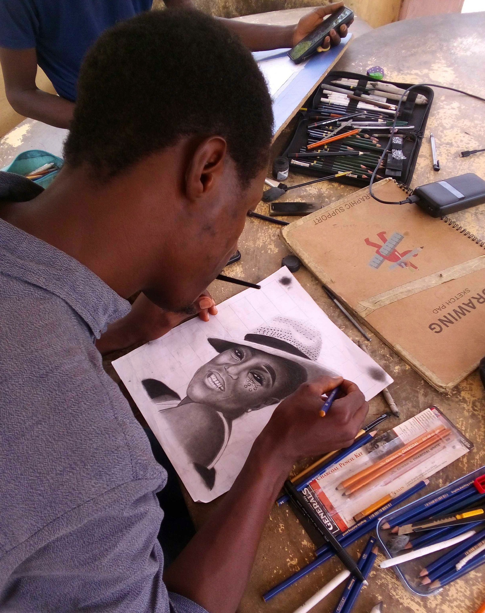 Person drawing a realistic portrait of a woman on paper, surrounded by art supplies and tools on a table.