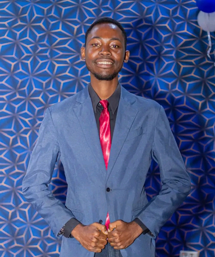 Young man in a suit with a red tie, smiling, standing against a geometric blue background.